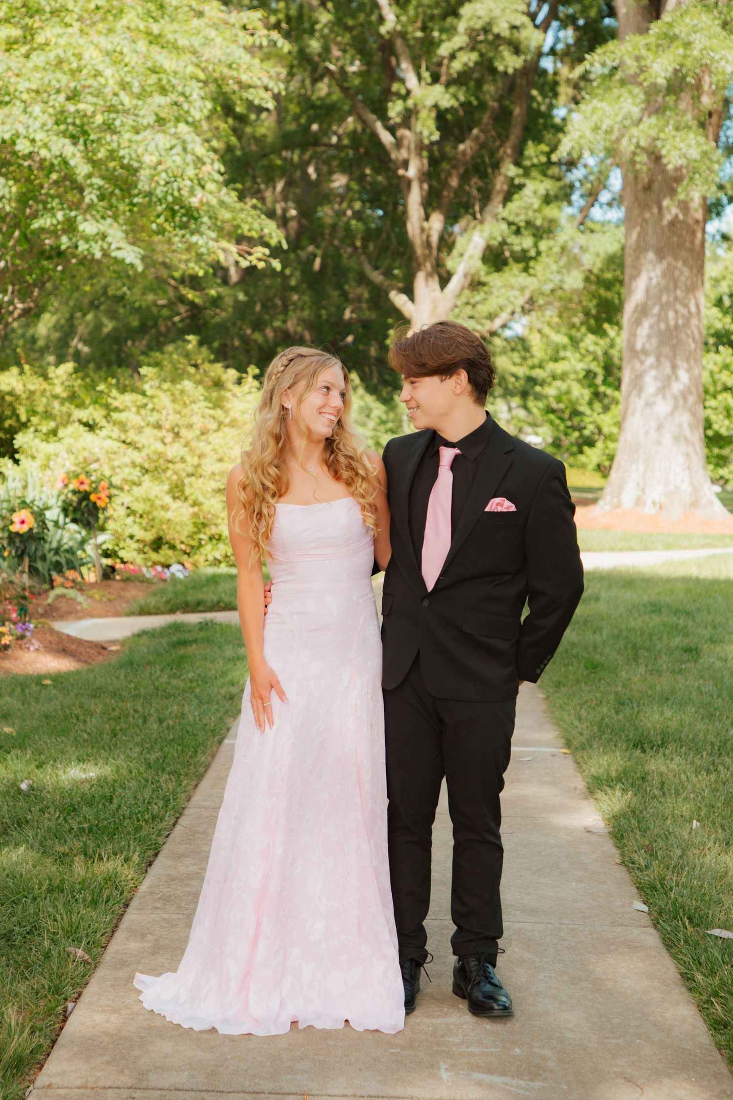 Teen prom couple in matching pink and black formal wear walk together on sunny garden path.