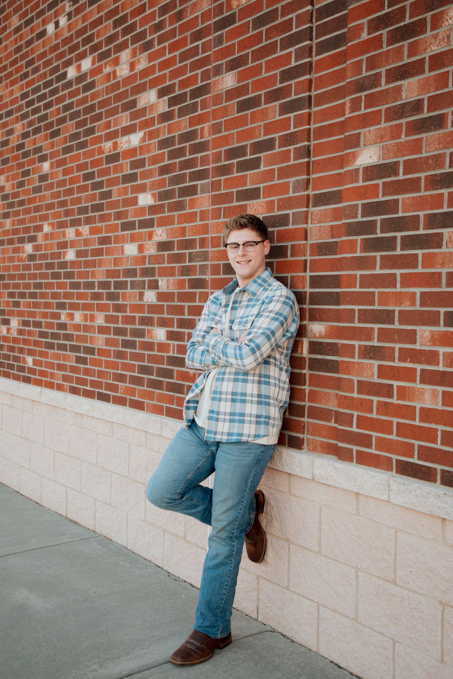 Teen boy in blue plaid shirt leaning against red brick wall with arms crossed.