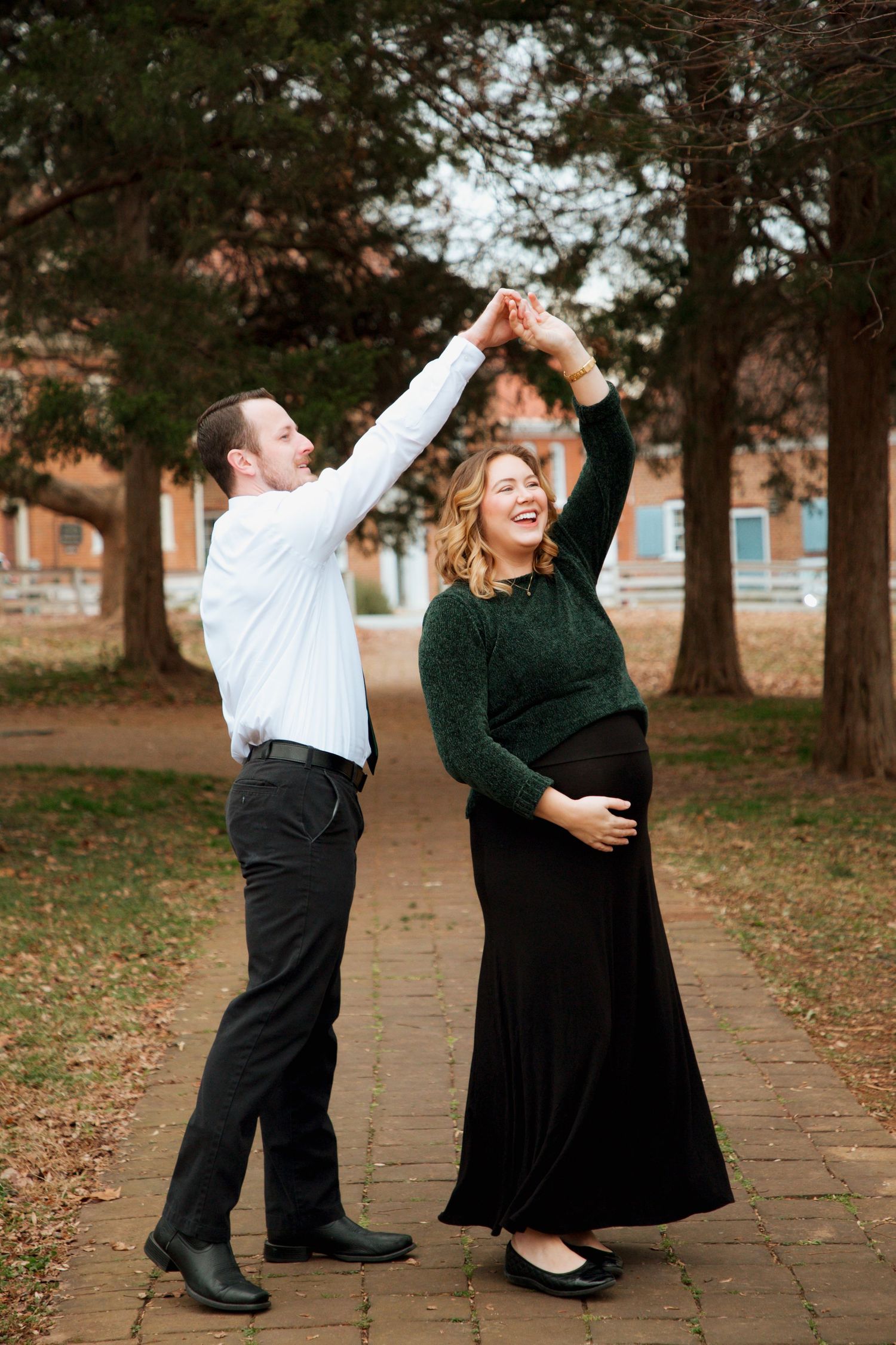 Laughing pregnant woman in green sweater dances with man in white shirt on brick pathway in autumn park.