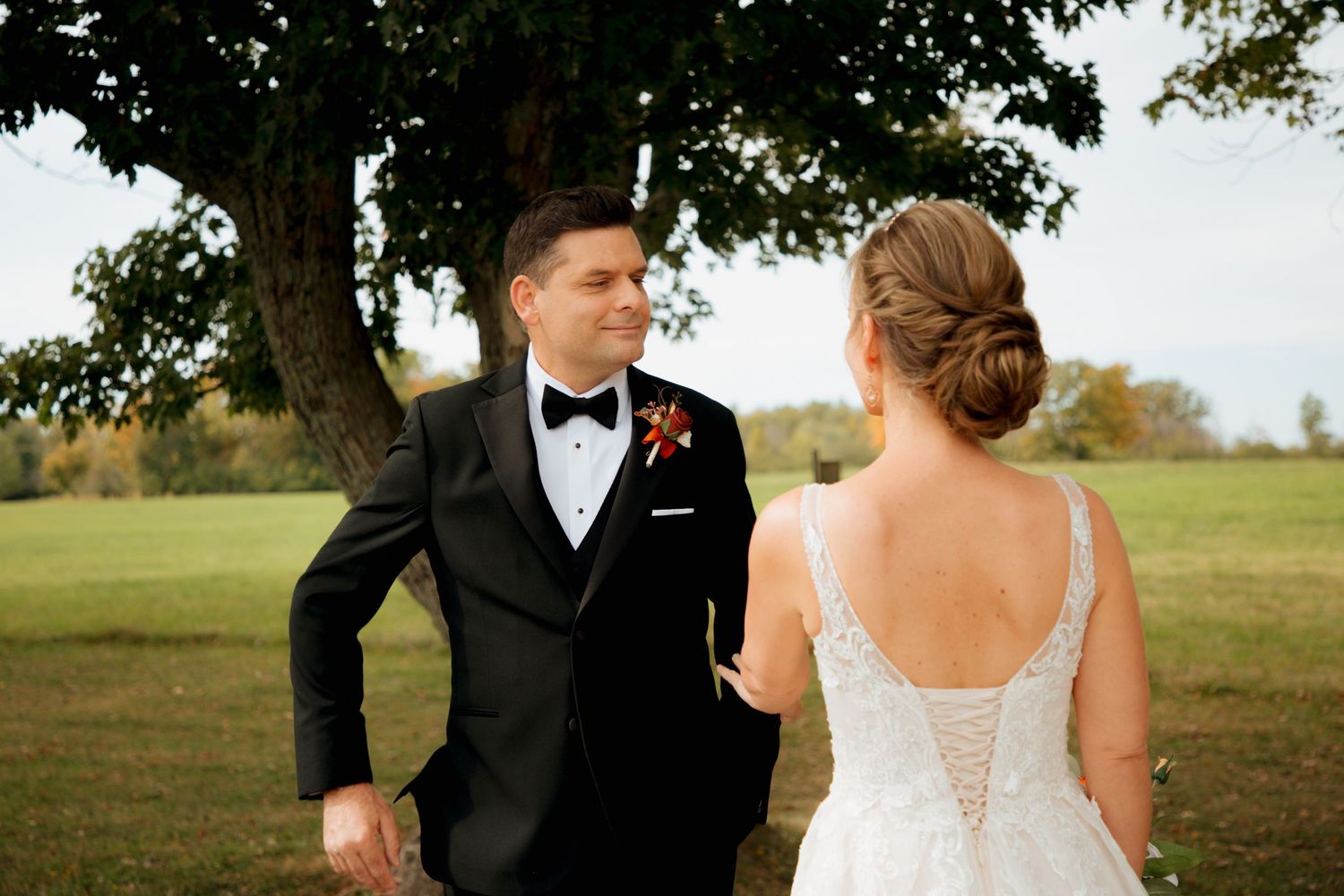 Groom in black tuxedo turns to see bride in white lace gown during romantic outdoor first look.