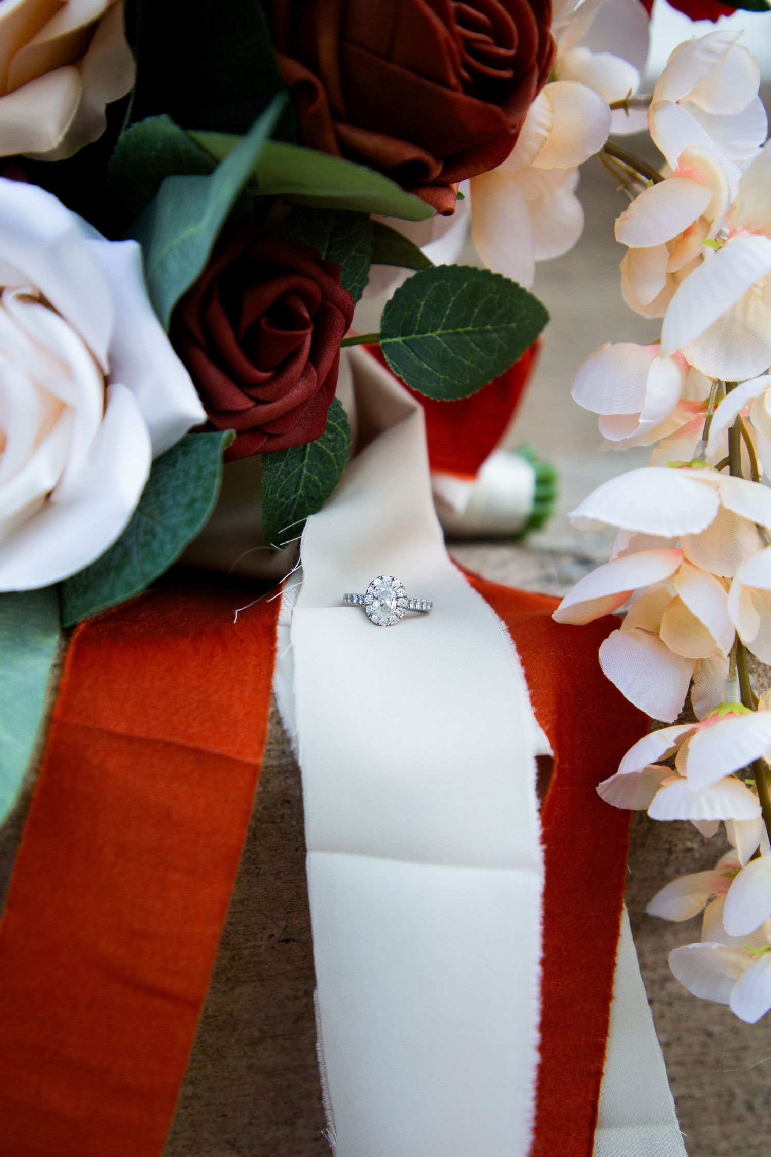 Diamond halo engagement ring displayed on white ribbon among red, white, and cream wedding bouquet flowers.