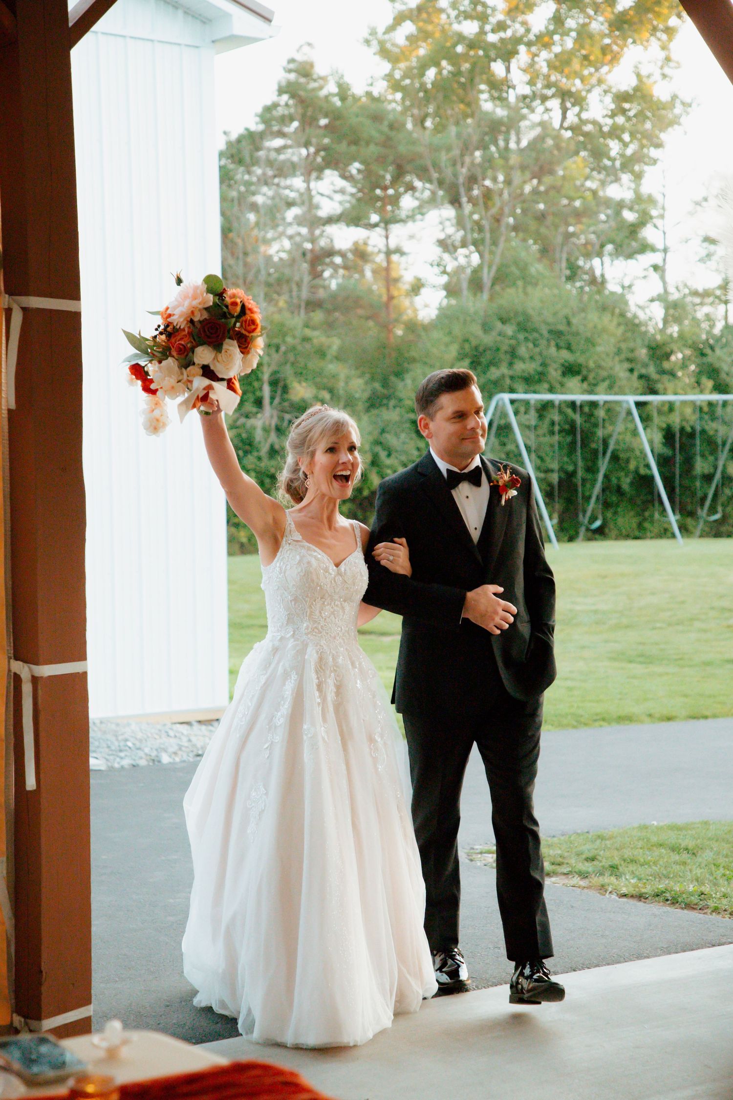 Joyful bride raises bouquet while groom smiles as they exit barn wedding venue.