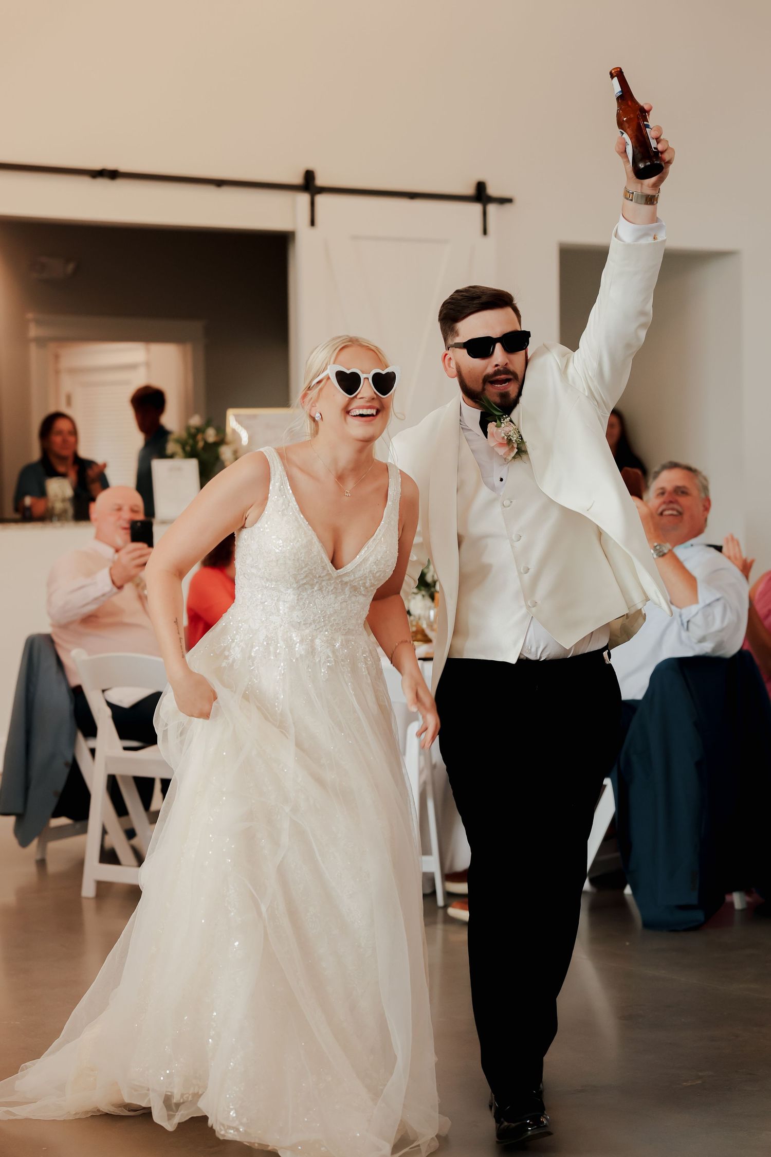 Bride and groom wearing heart sunglasses making grand entrance at wedding reception, groom raising beer bottle in celebration.