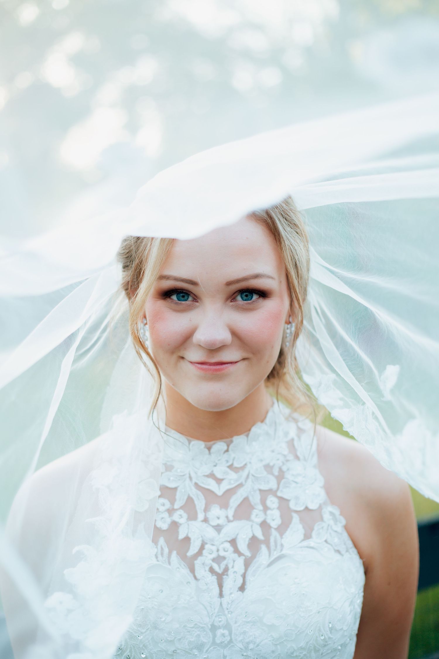 Beautiful blonde bride smiles softly under flowing white veil in lace gown.