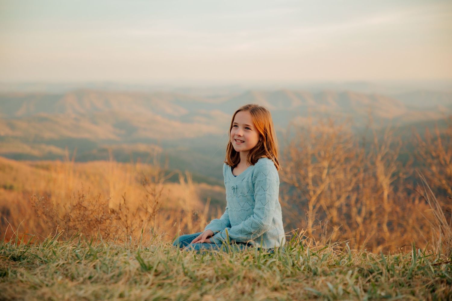 Smiling young girl sitting on a hillside with a scenic mountain landscape at golden hour.