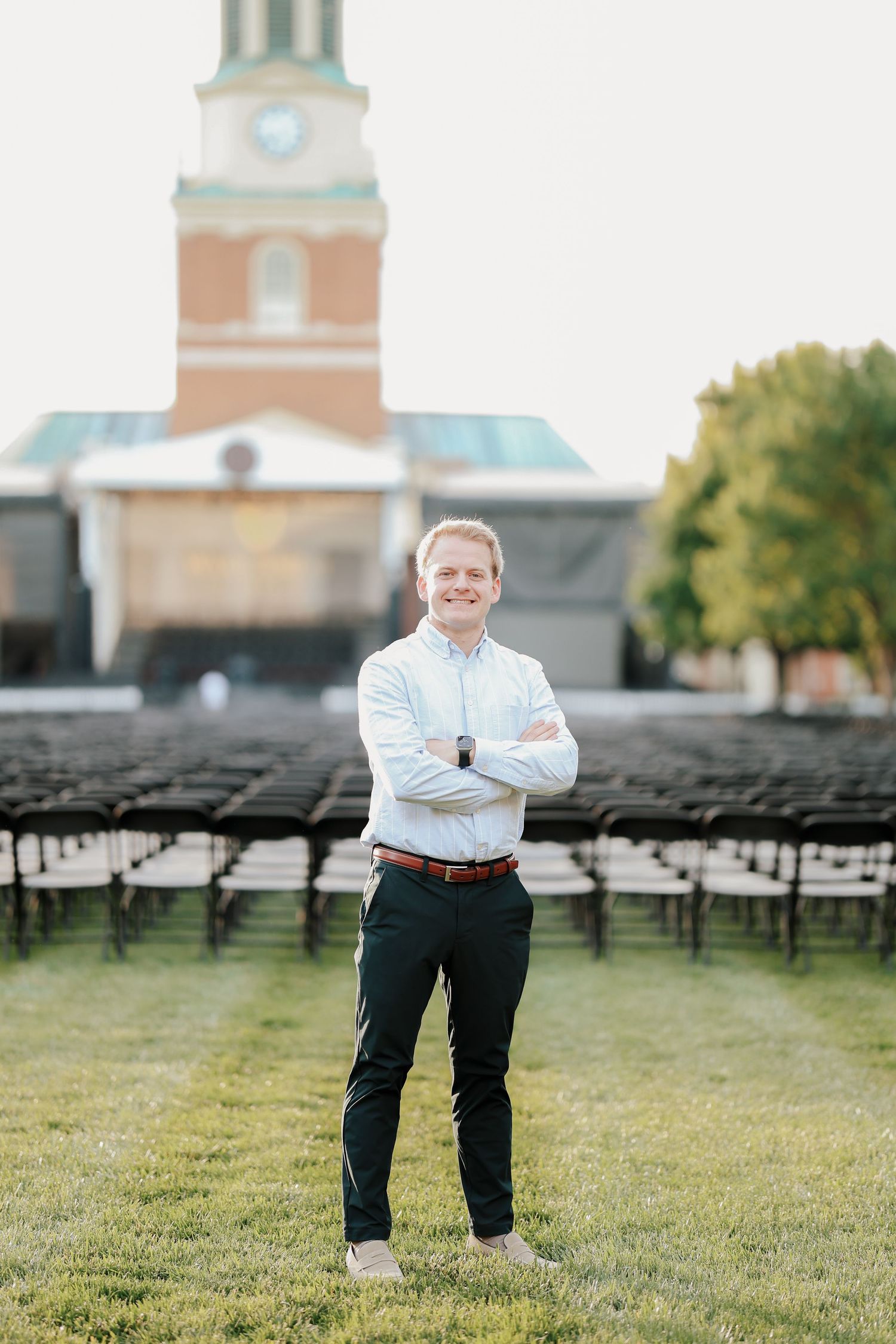 Smiling young man with arms crossed standing on green lawn with brick clock tower building behind him.