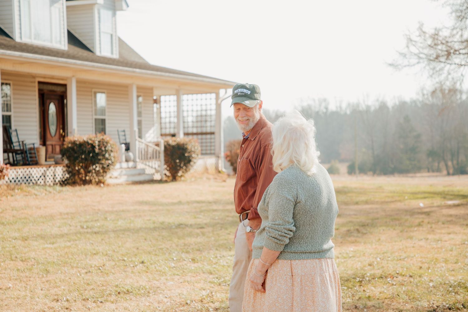 Elderly couple holding hands and sharing a quiet moment in front of a white farmhouse.