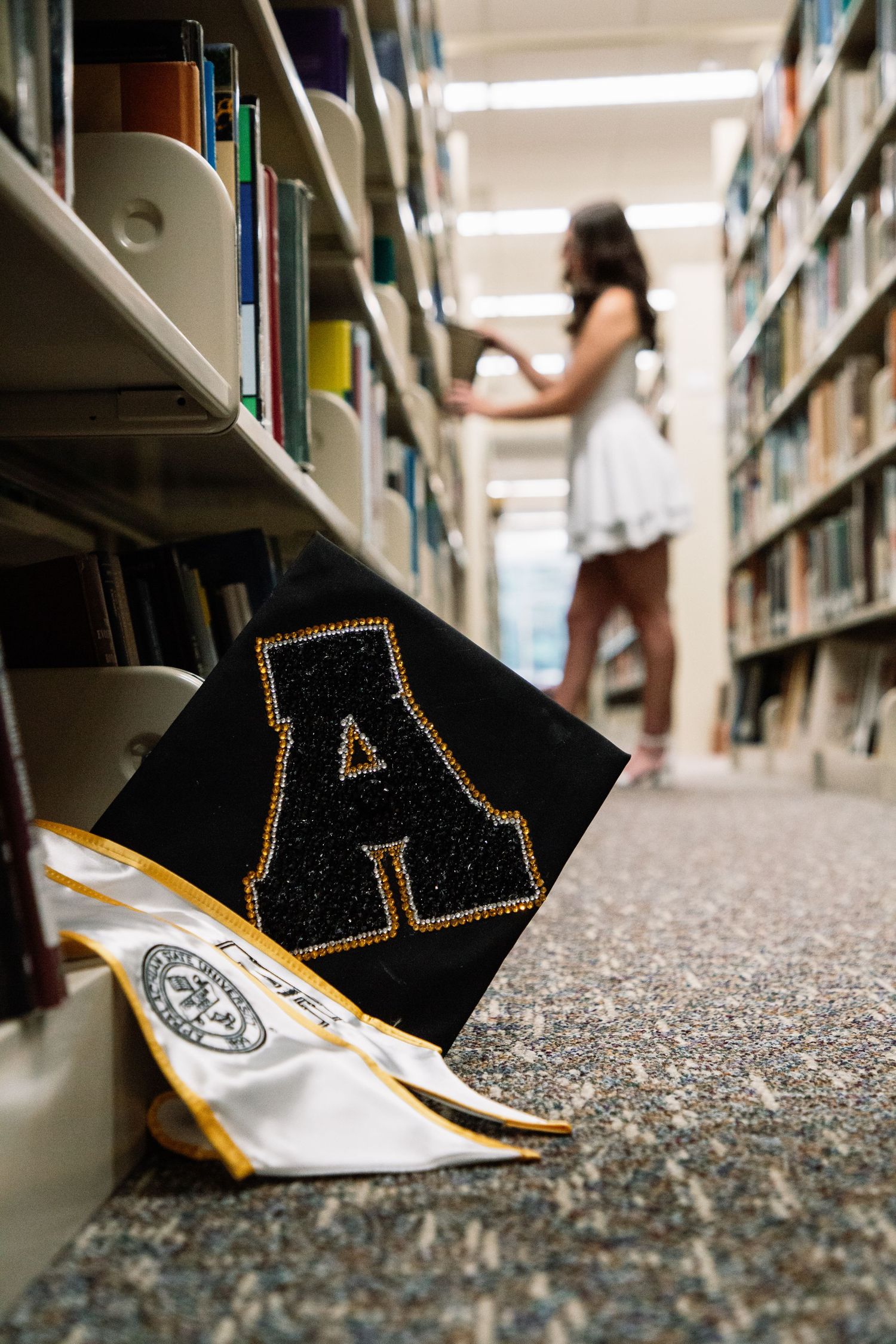 Graduation cap with gold letter A and white stole on library floor with graduate browsing books.
