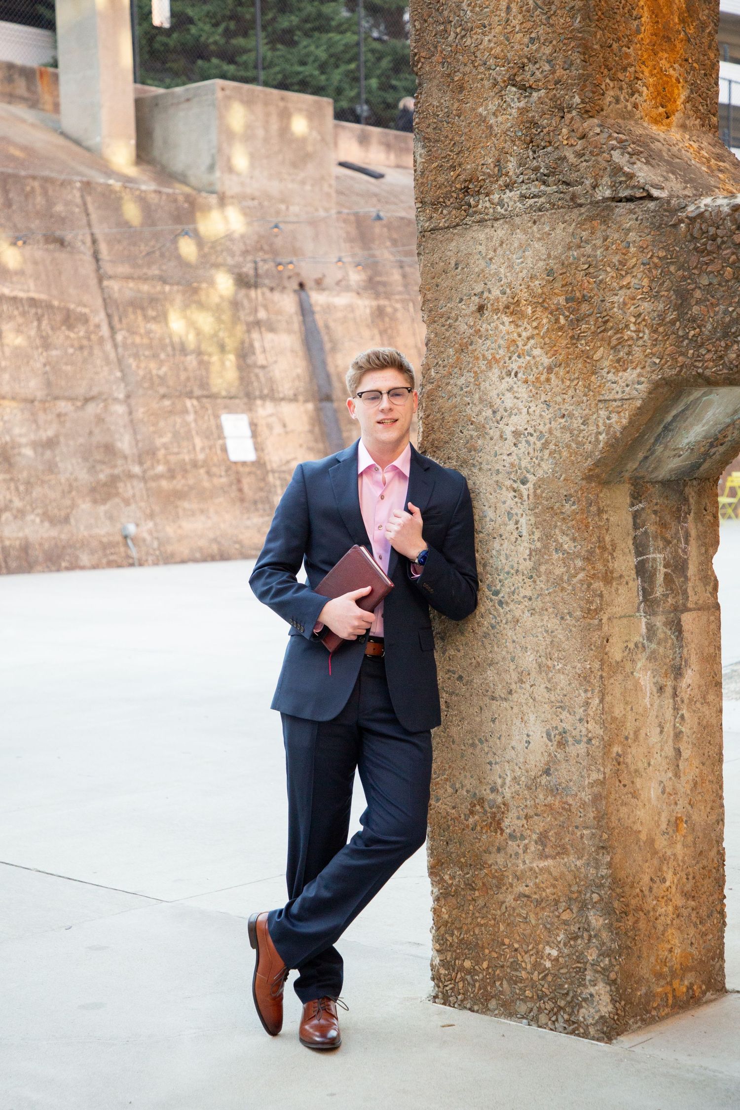 Young man in navy suit holding Holy Bible with graduation tassel poses against stone column outdoors.