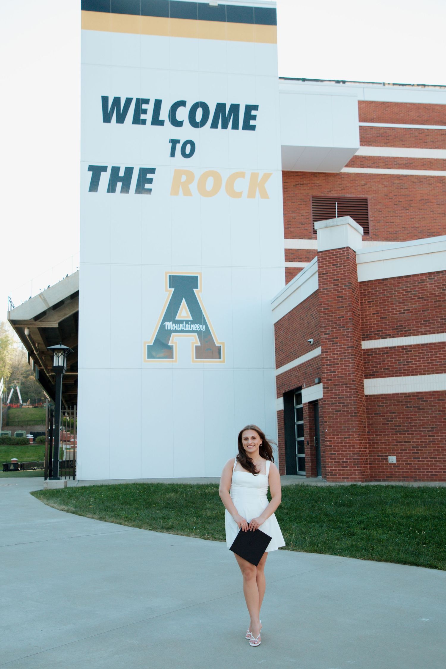Female graduate in white dress posing at Welcome to The Rock Appalachian State stadium.