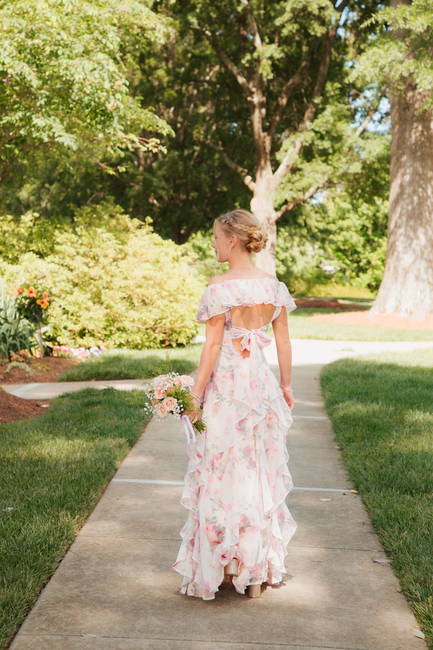 Young woman in pink floral off-shoulder ruffle gown walking away on garden path holding small bouquet.