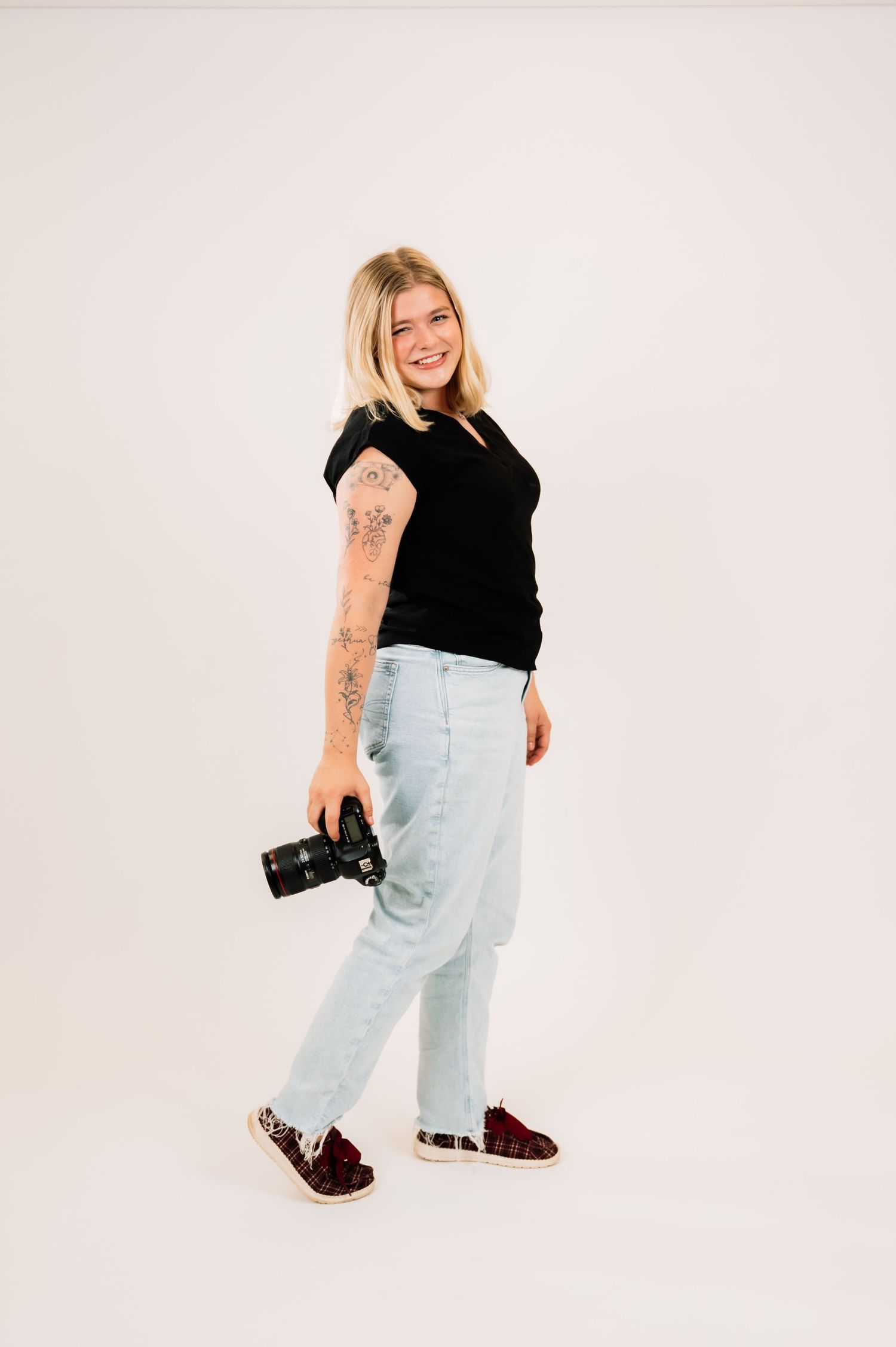 Tattooed blonde woman walking in profile holding DSLR camera wearing black tee and light wash jeans.