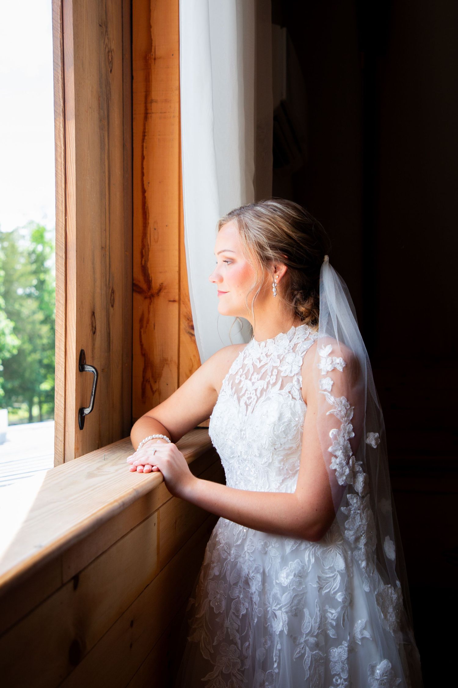 Bride in lace wedding dress and veil gazes out rustic wooden window in soft natural lighting.
