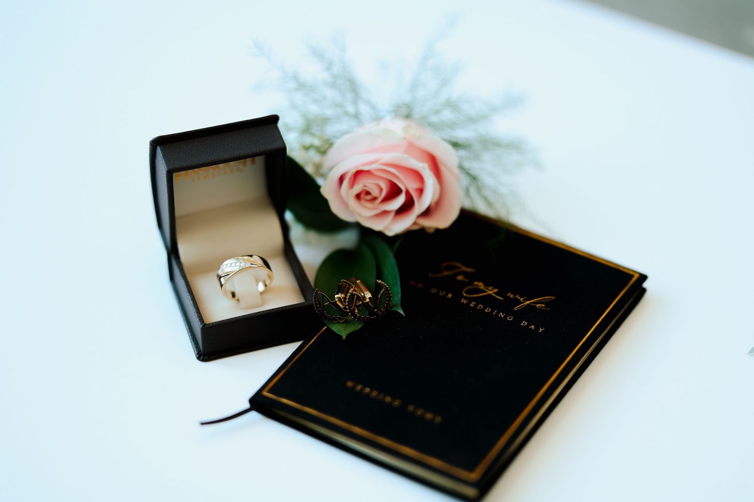 Wedding ring in black box beside pink rose and black vow book on white surface.