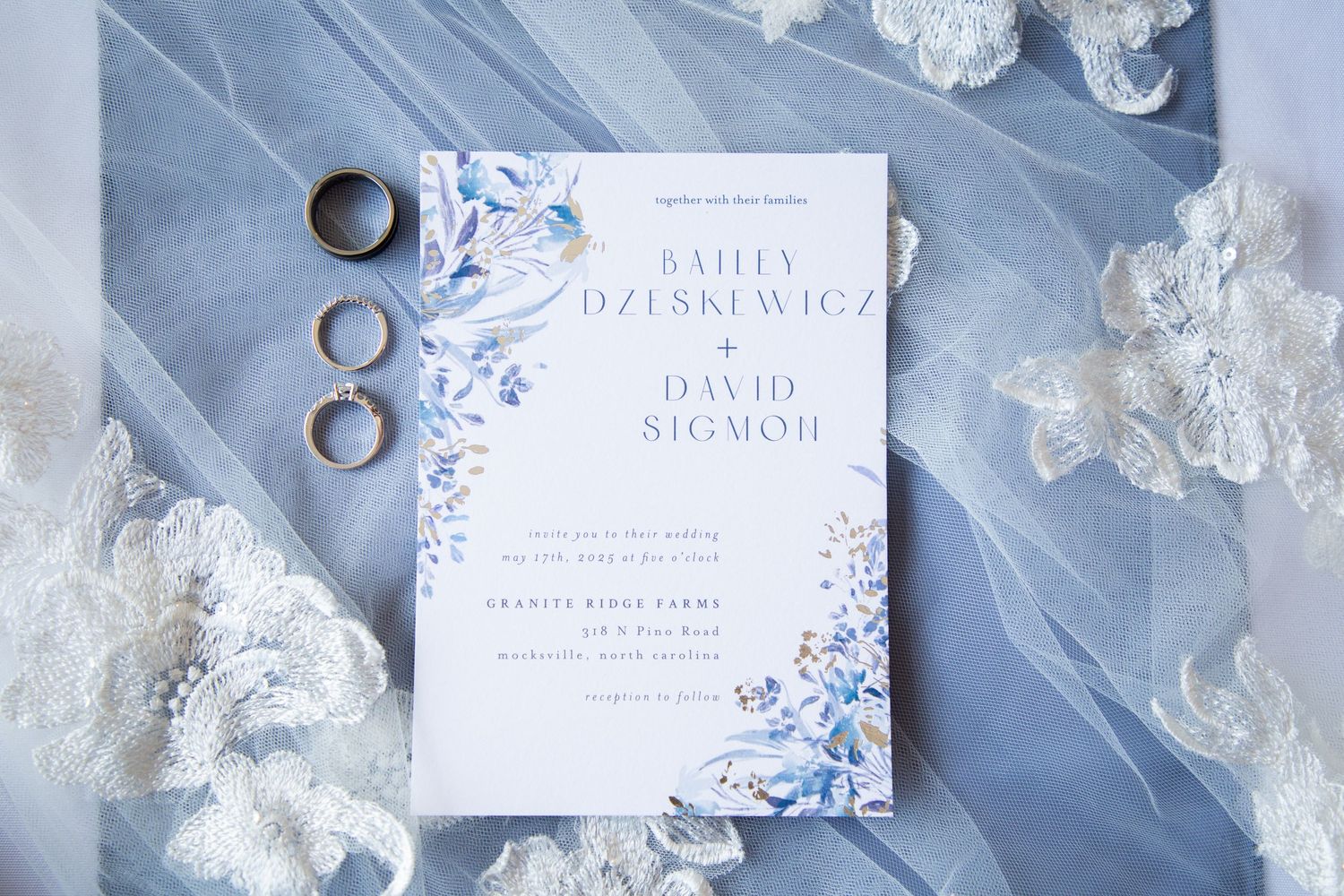 Blue floral wedding invitation for Bailey Dzeskewicz and David Sigmon with gold rings on dusty blue fabric.