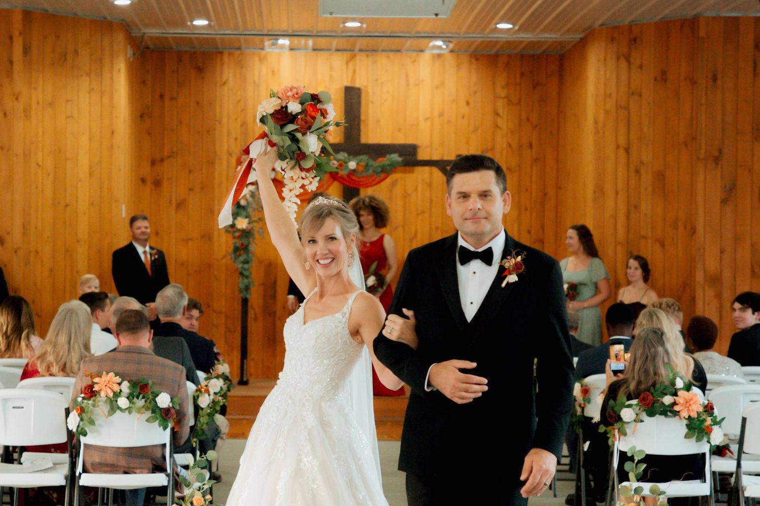 Joyful bride raises bouquet while walking down the aisle with groom after wedding ceremony in rustic chapel.