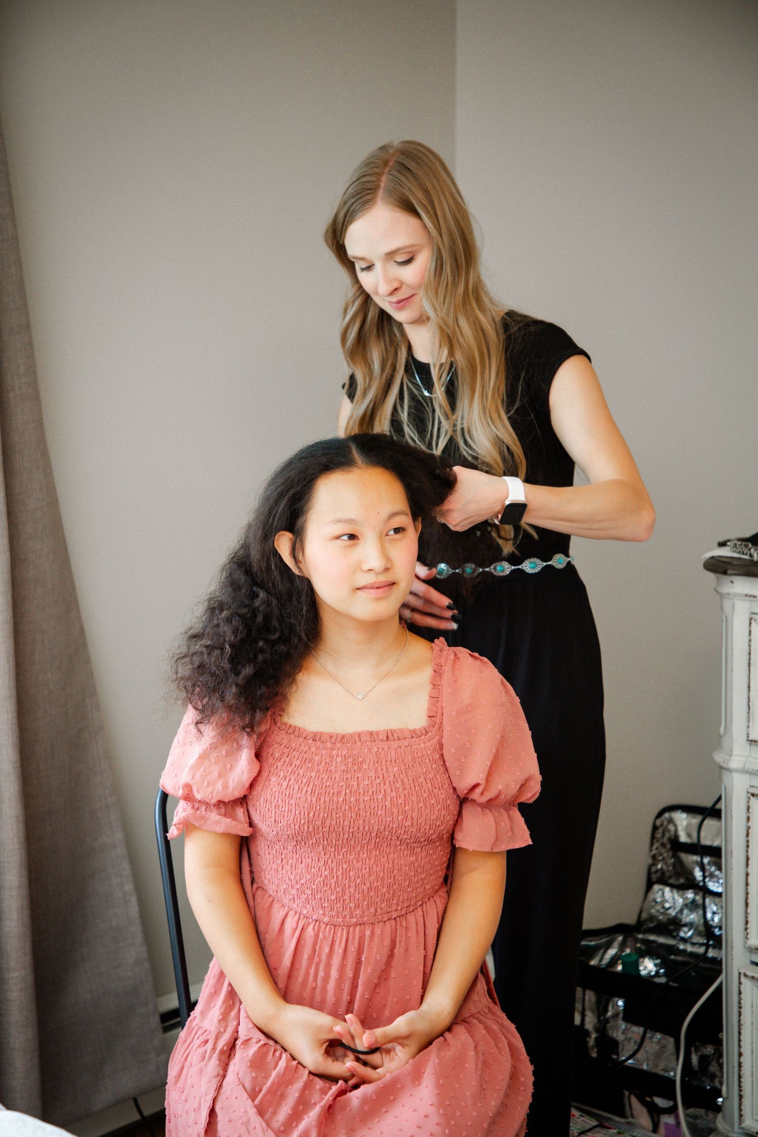 Hairstylist in black dress styling young girl with curly hair in pink dress.