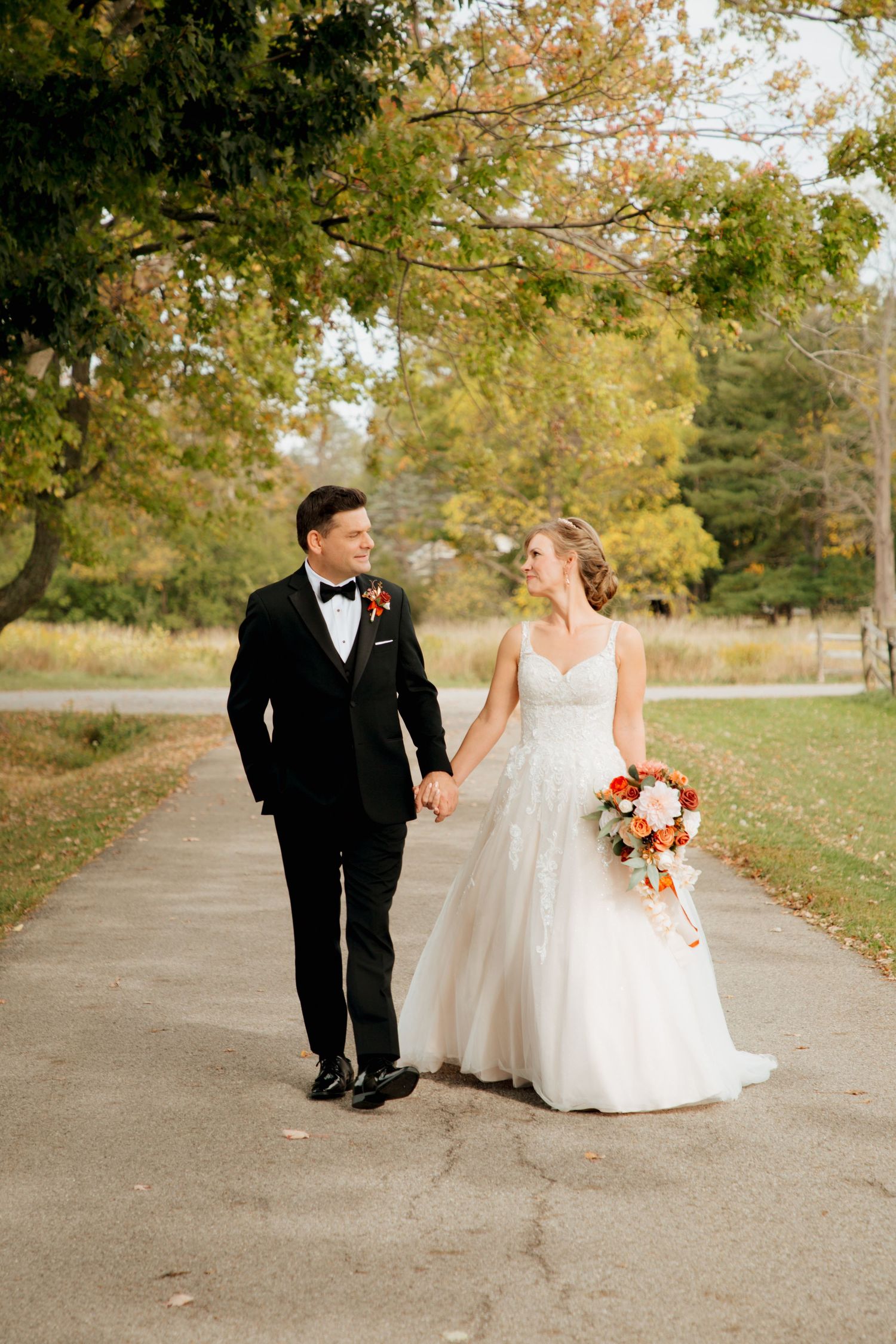 Smiling bride and groom walk together on fall path surrounded by golden autumn trees.