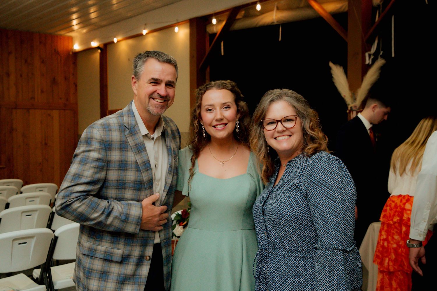 Three guests smile together at a wedding reception in a rustic venue.