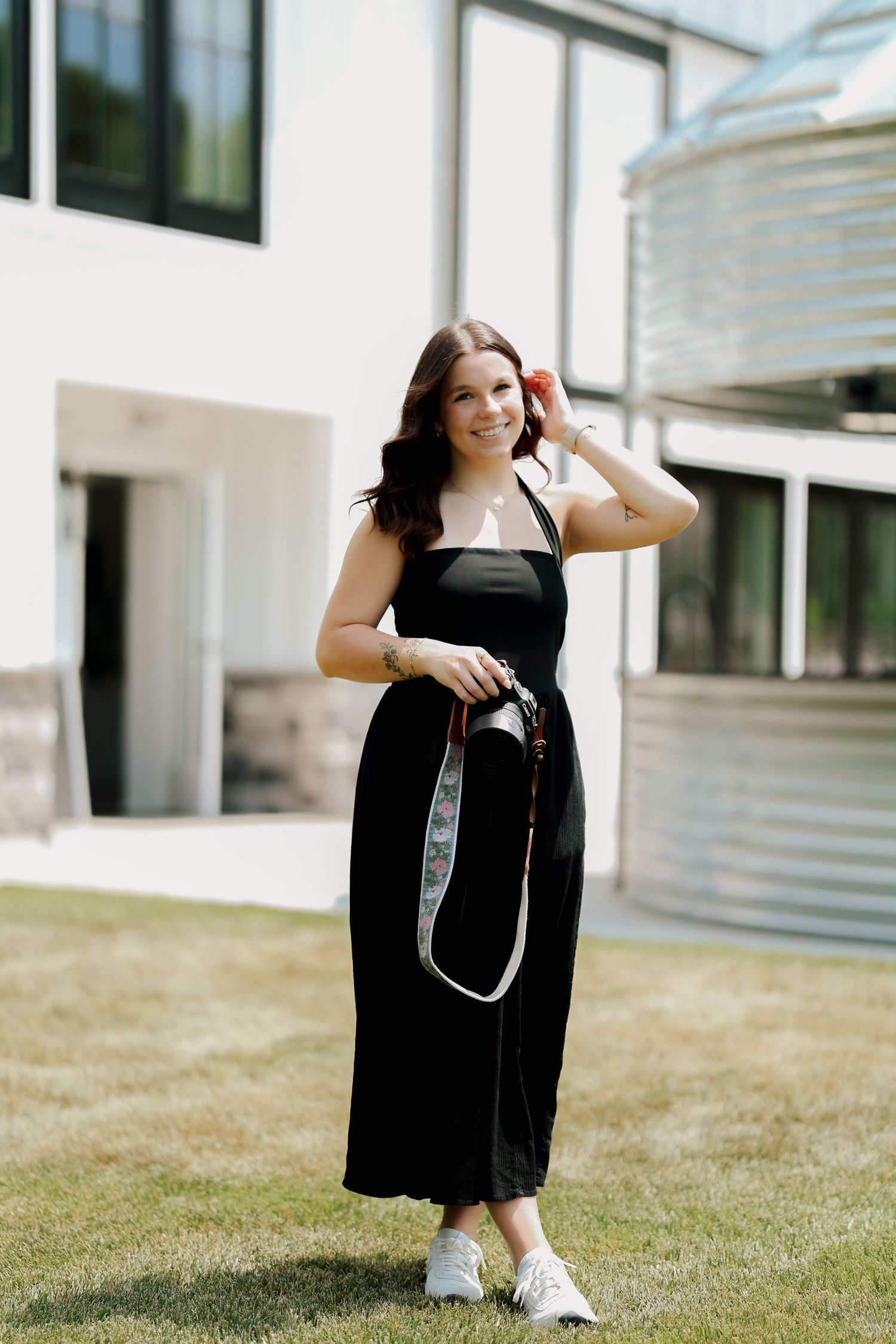 Young woman in black midi dress holding camera strap, posing outdoors near modern white building on green lawn.