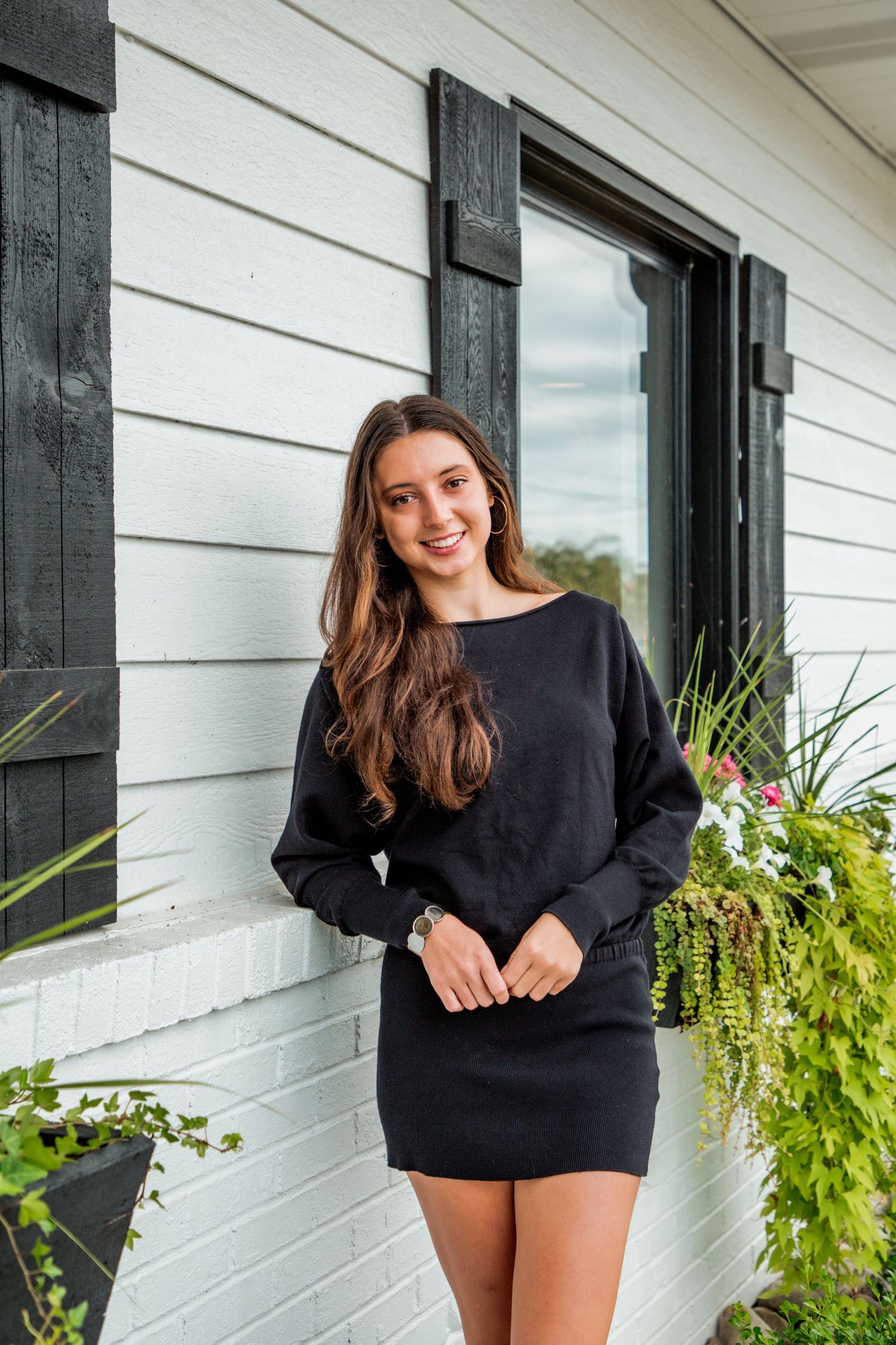 Smiling young woman in black mini dress standing on porch with flower boxes beside white house.