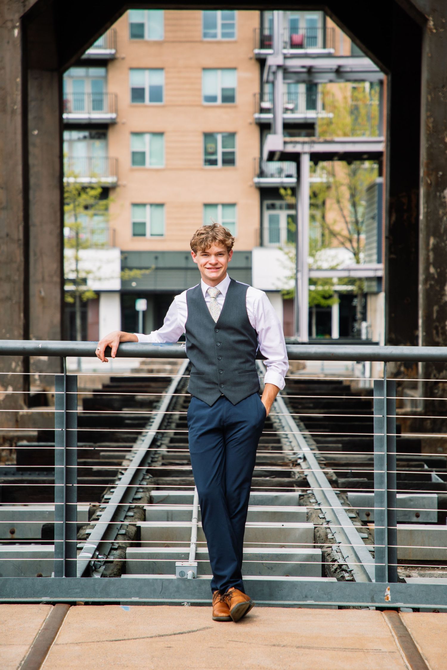 Smiling young man in navy suit vest posing on railroad tracks beneath urban industrial bridge.