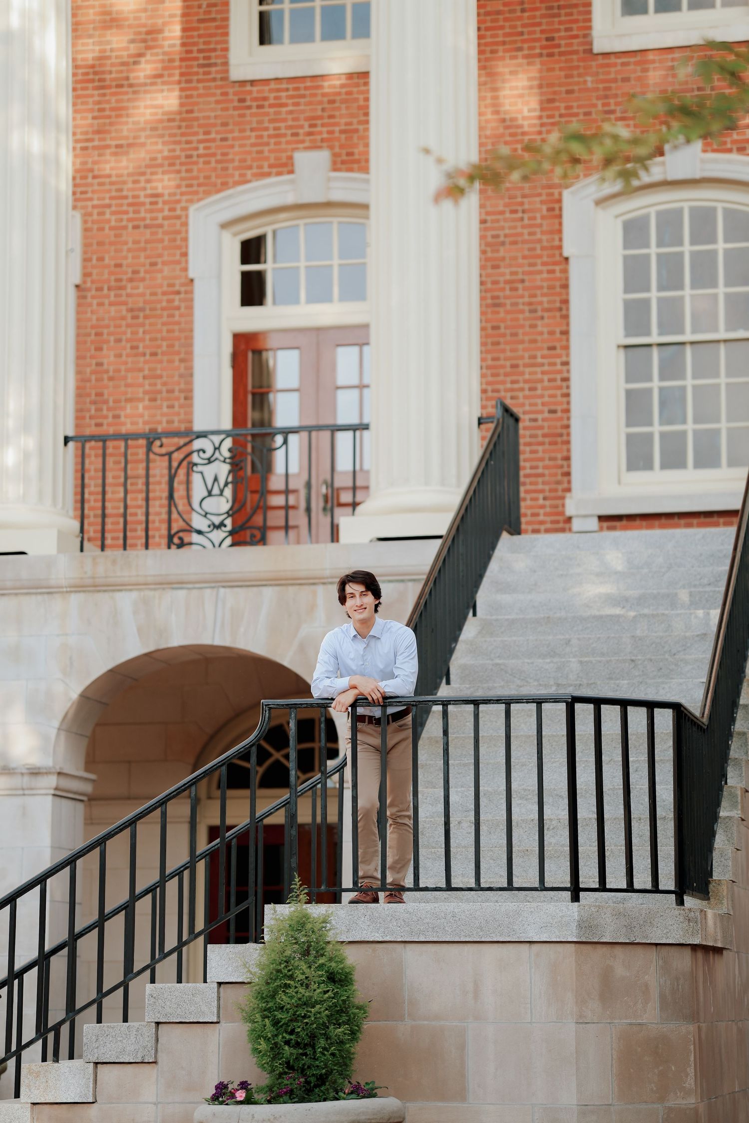Smiling young man in blue shirt leans on black railing on the steps of a brick campus building.