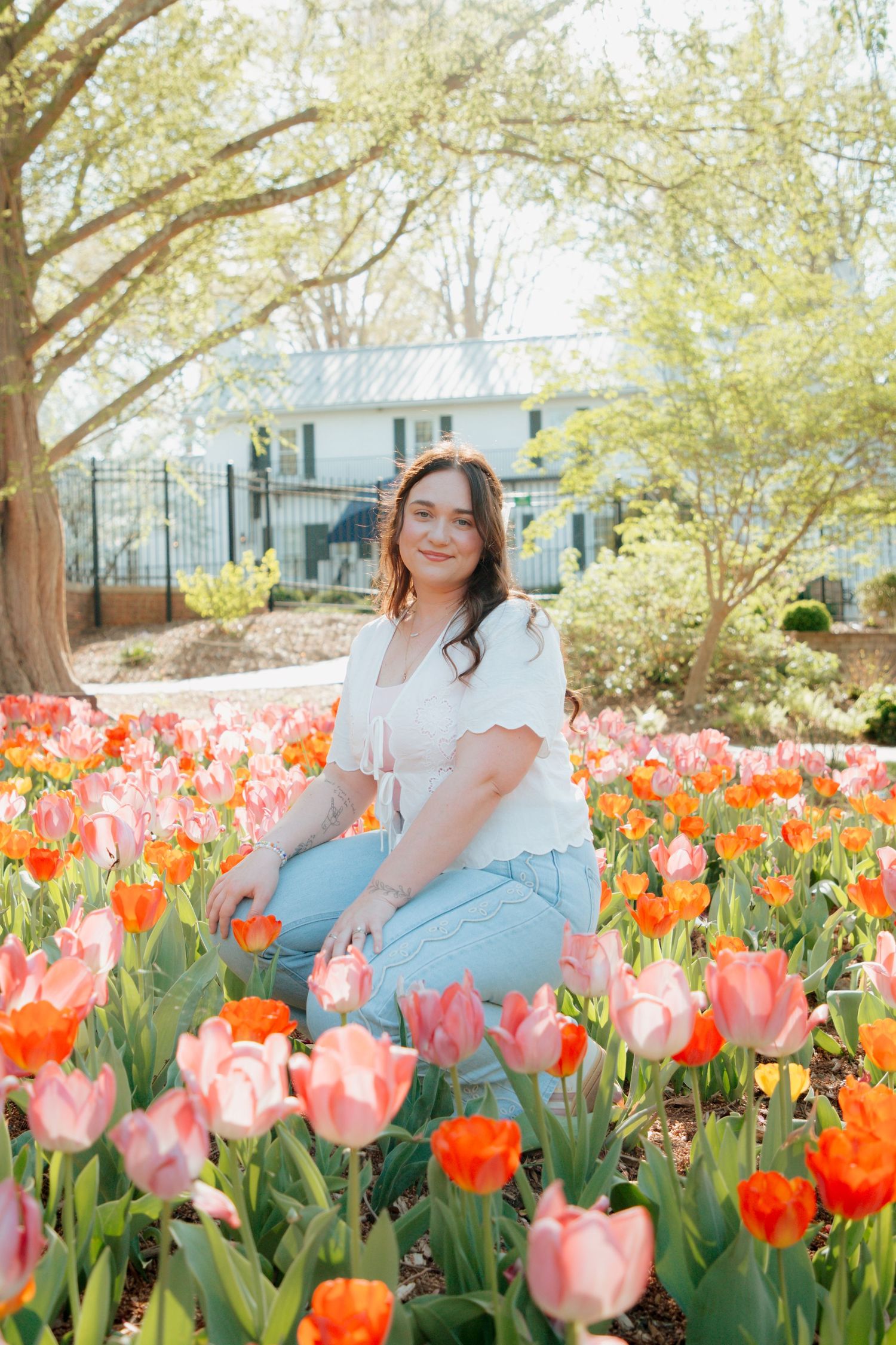 Brunette woman sits smiling among pink and orange tulips in a sunlit spring garden.