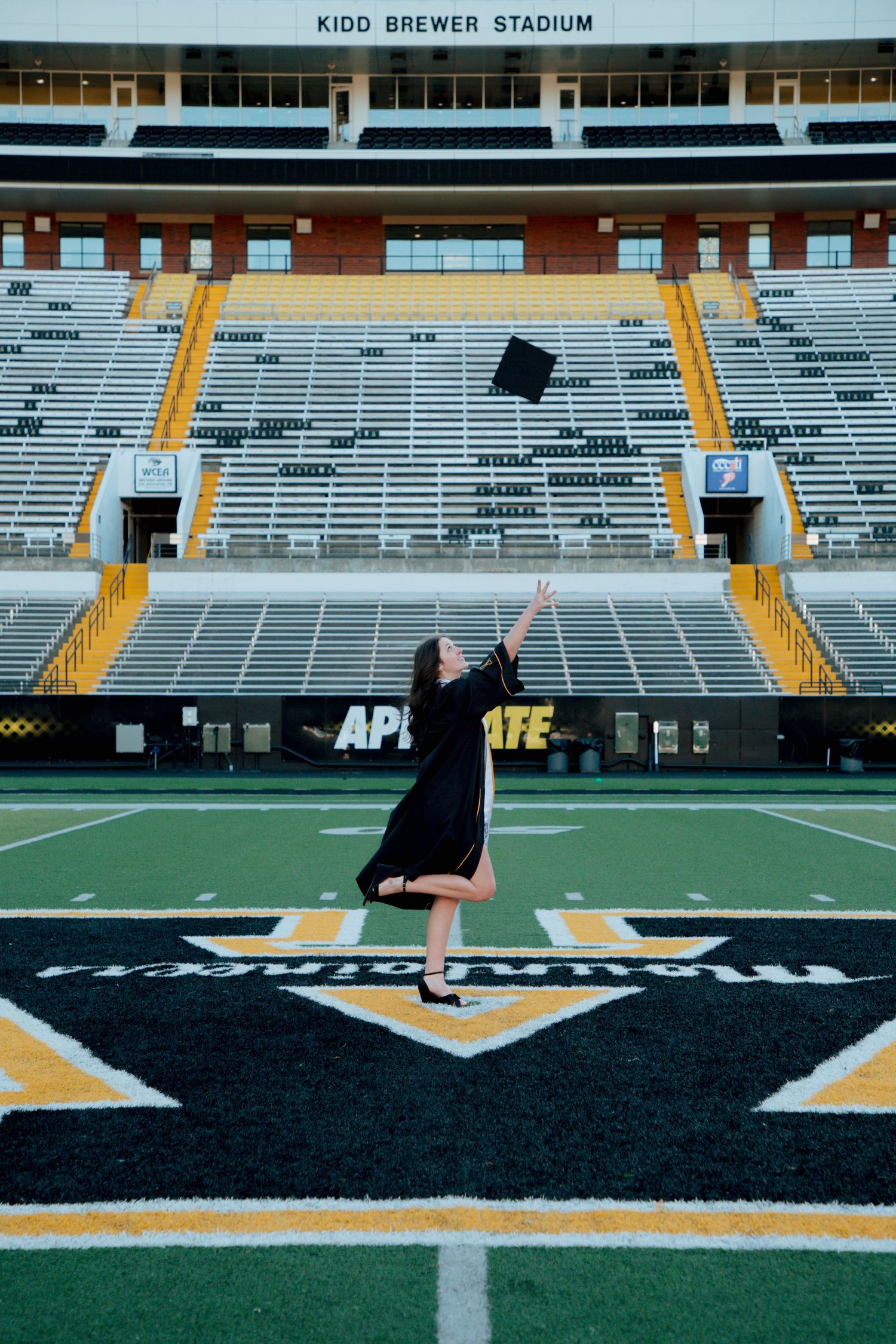 Graduate in black gown tosses cap at Kidd Brewer Stadium, Appalachian State University football field.