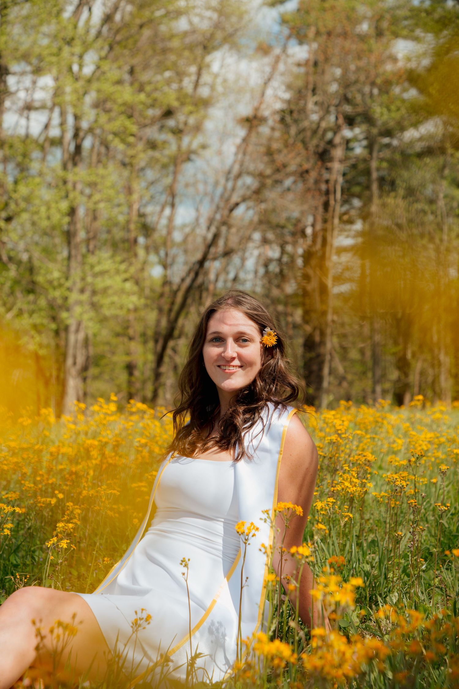 Smiling female graduate in white dress sitting among yellow wildflowers with flower in her hair.