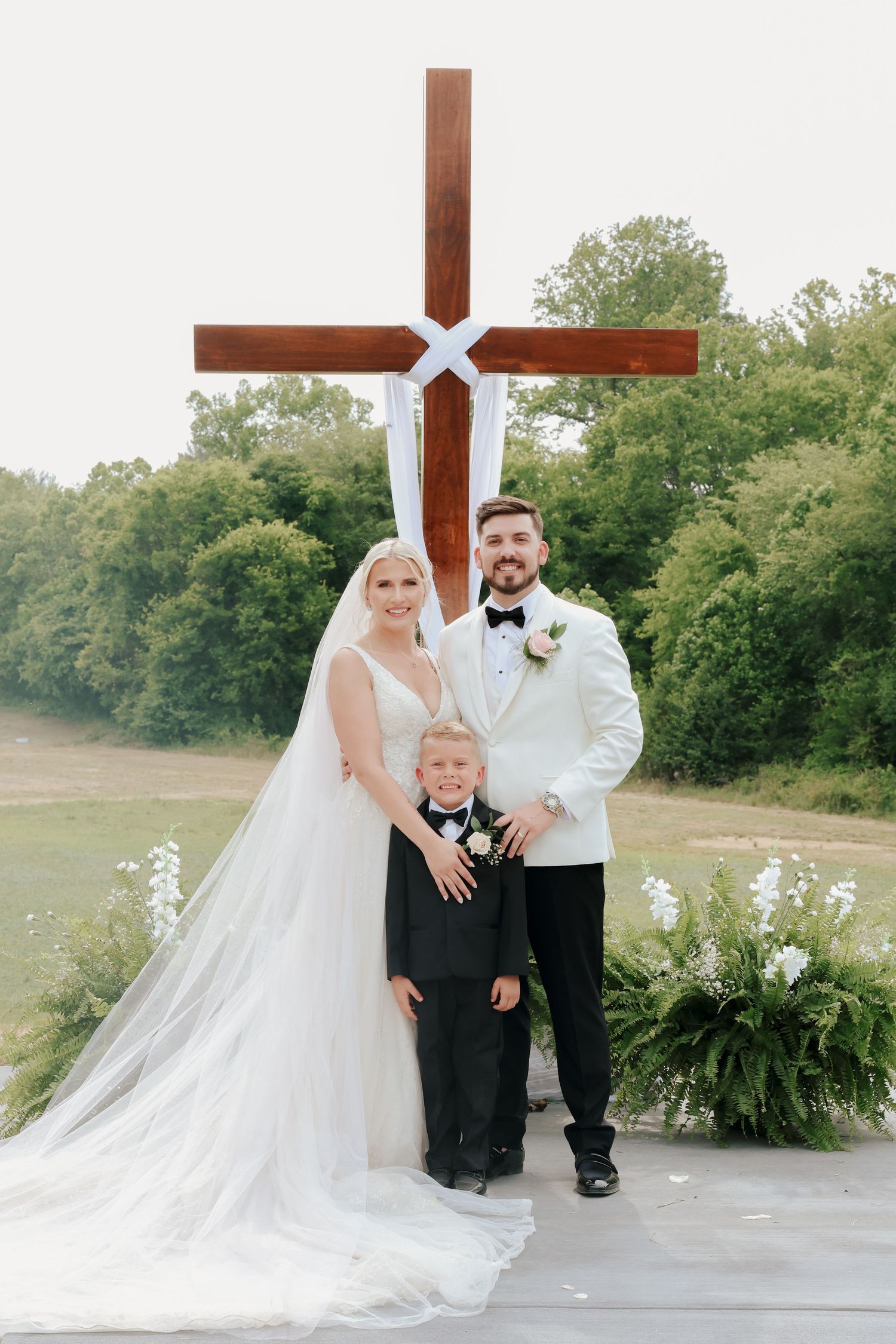 Bride and groom posing with young boy in tuxedo before large wooden cross at outdoor wedding ceremony venue.