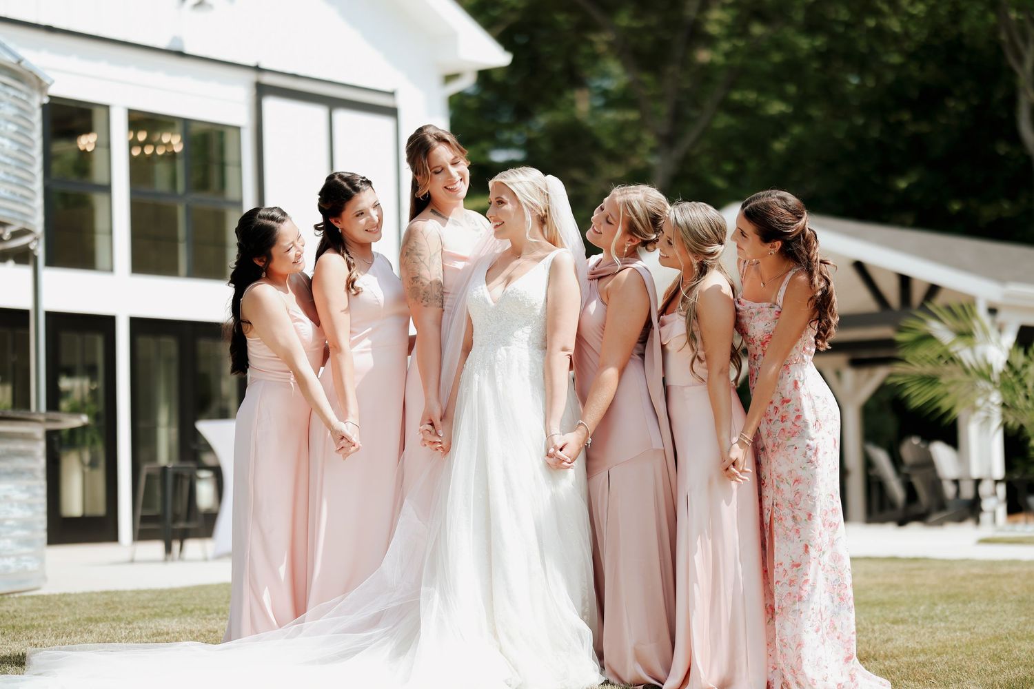 Bride surrounded by bridesmaids in blush pink gowns sharing a joyful moment outside a white venue.