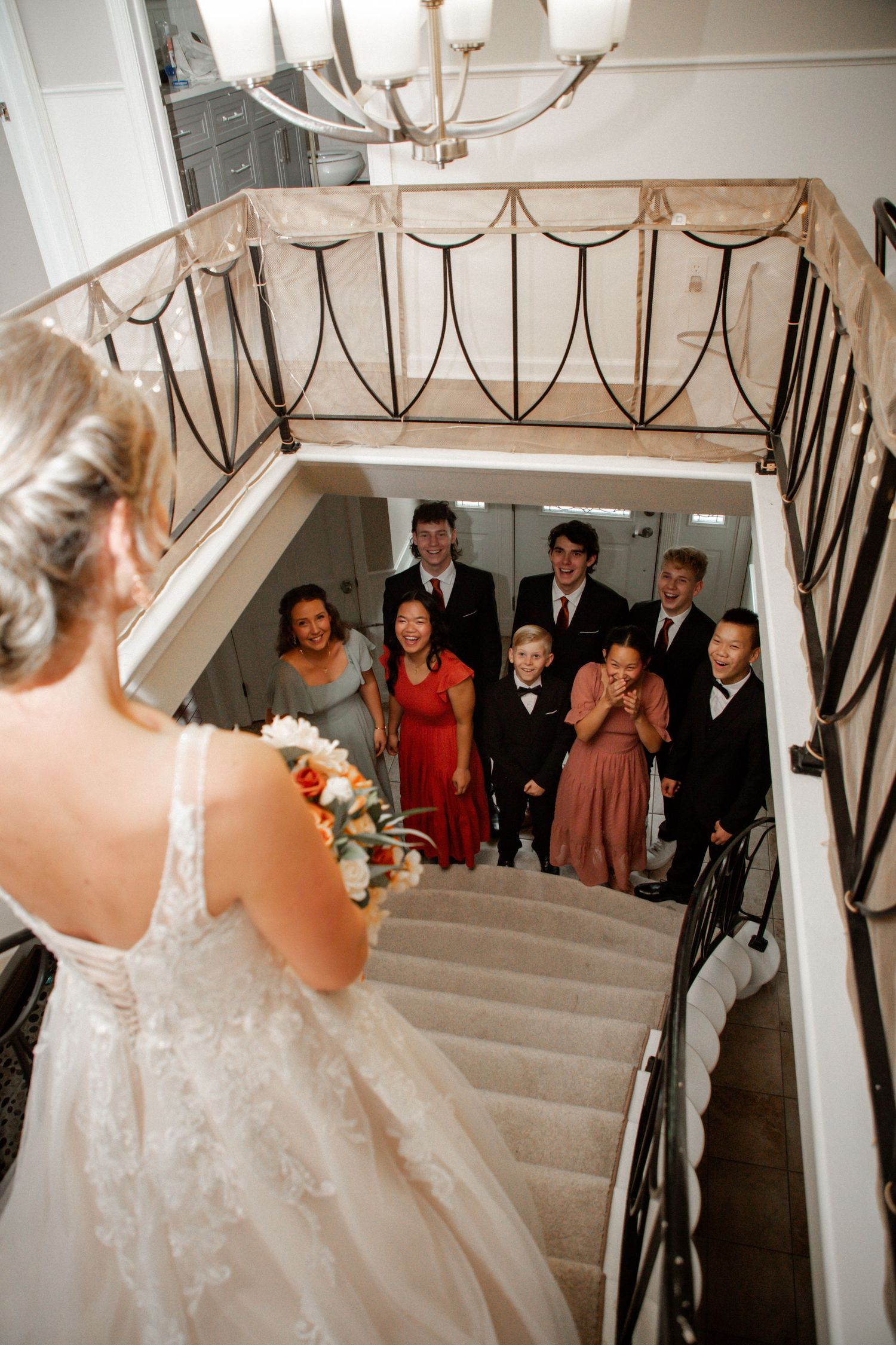 Bride descends grand staircase in white lace gown as smiling wedding party gathers below to greet her.