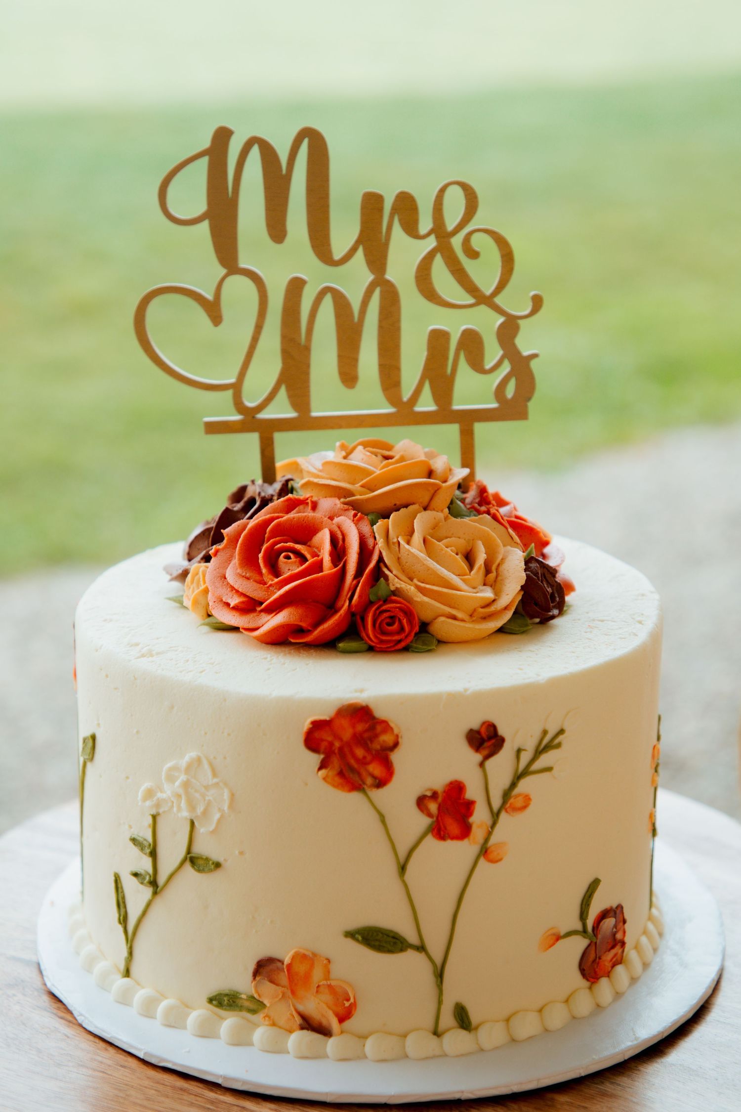 White wedding cake with hand-painted floral design, orange and red buttercream roses, and gold Mr and Mrs cake topper.