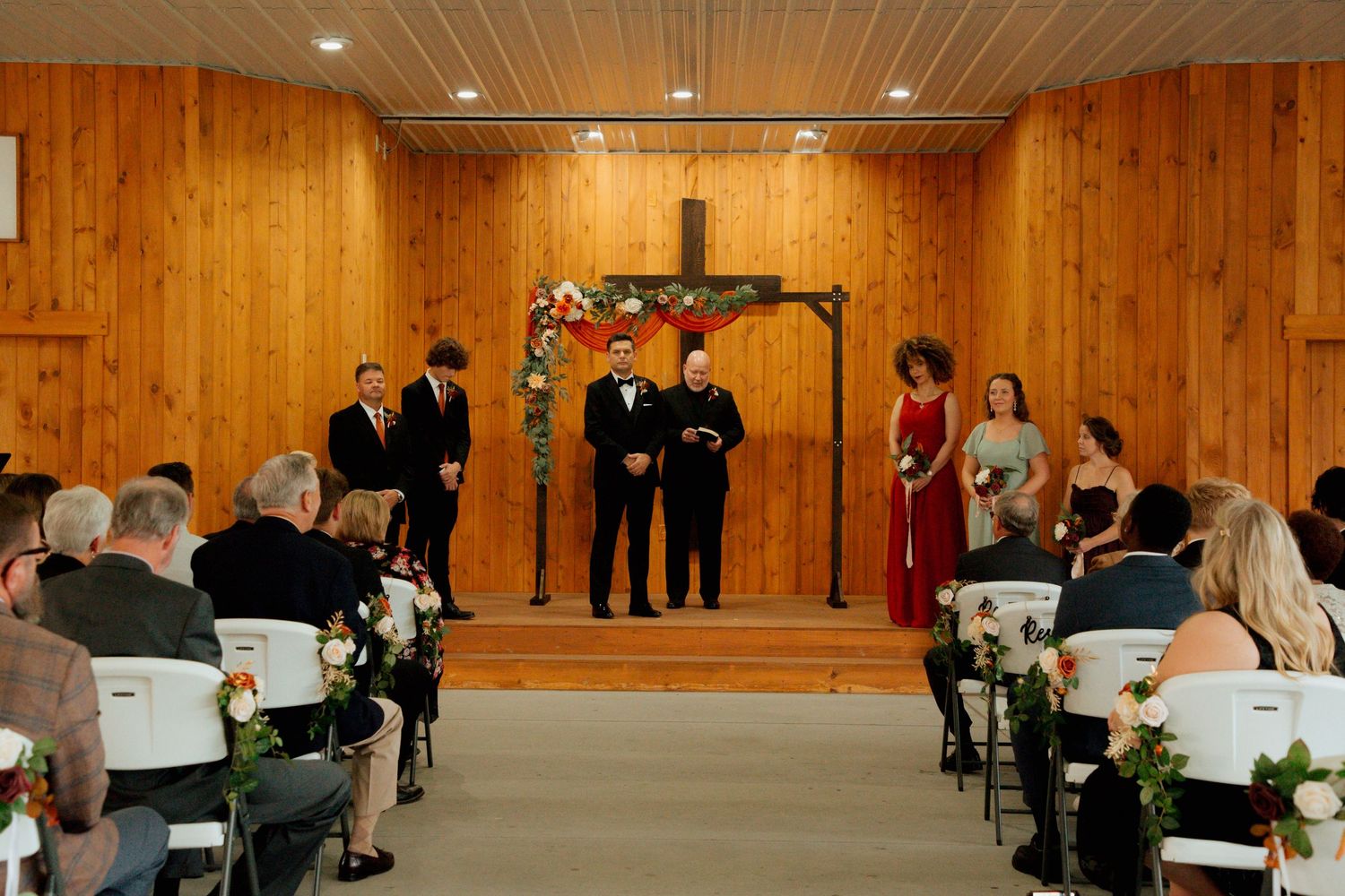 Wedding ceremony inside a wood-paneled chapel with guests seated as groom waits at floral arch.