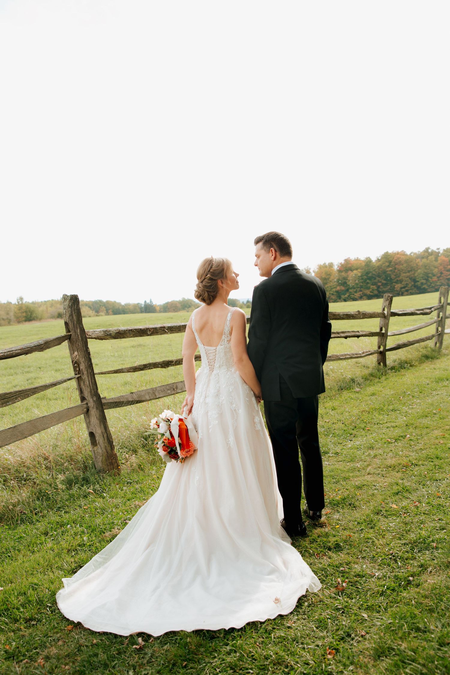 Bride and groom standing by rustic fence overlooking green pasture.