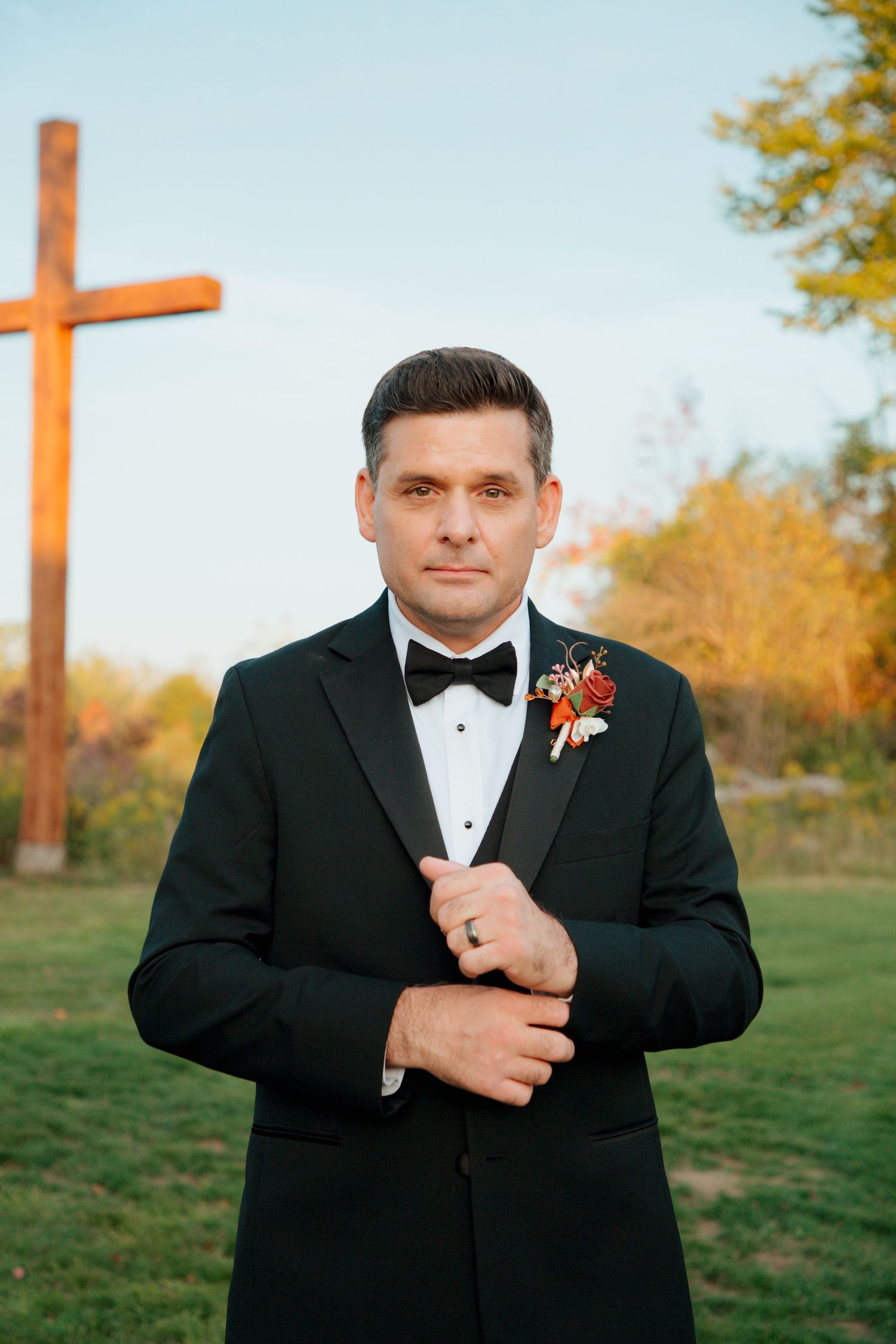 Groom in black tuxedo with boutonniere poses before wooden cross in autumn landscape.