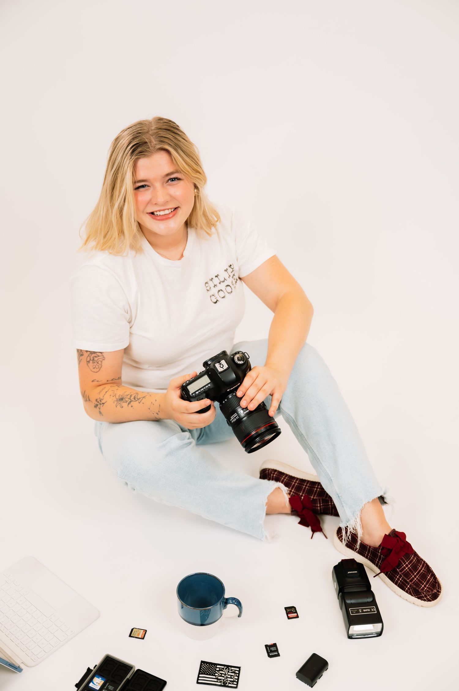 Smiling blonde photographer sitting on floor holding DSLR camera surrounded by photography equipment and accessories.
