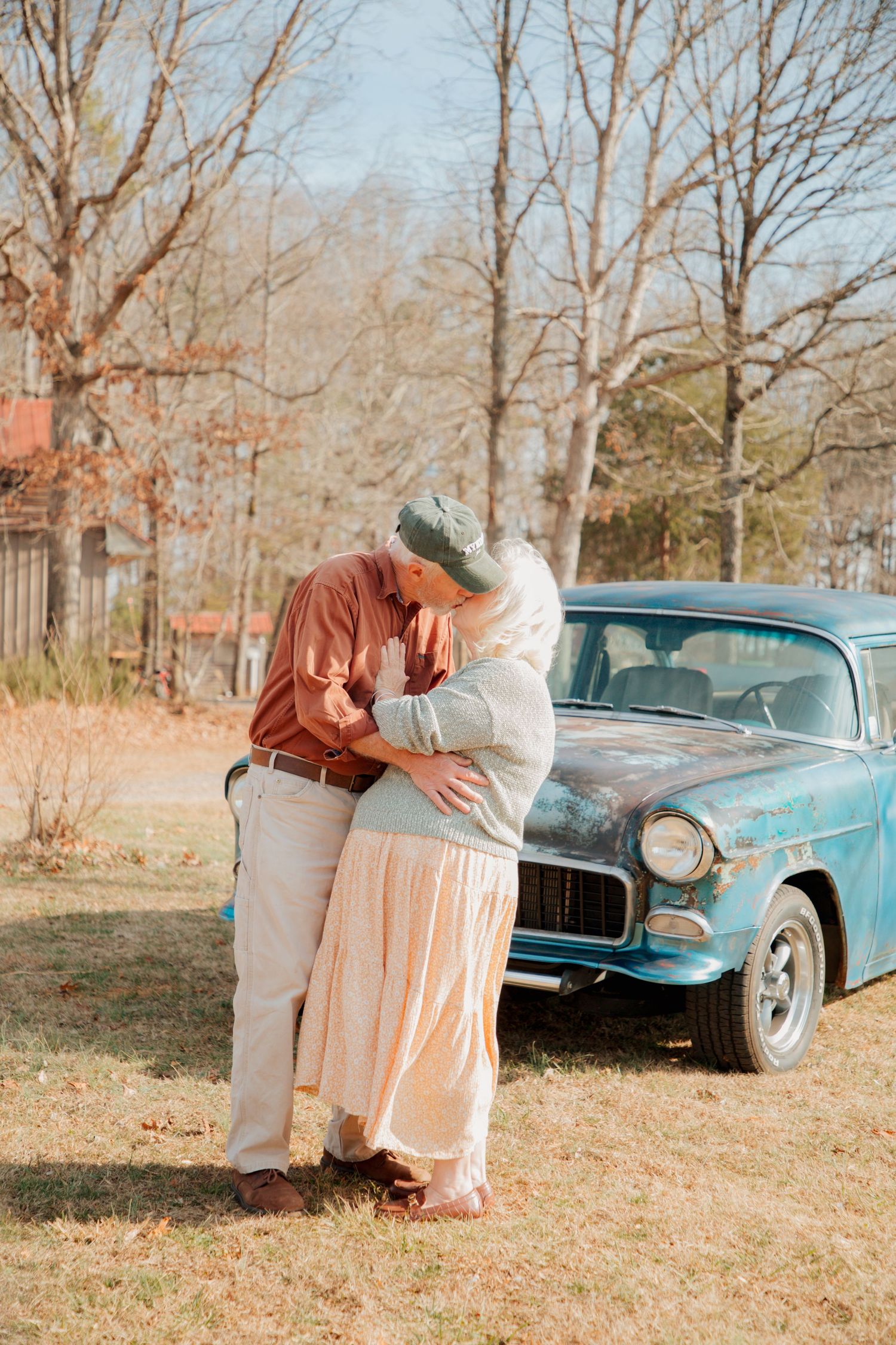 Elderly couple sharing a romantic kiss beside a vintage teal classic car on a sunny autumn day.