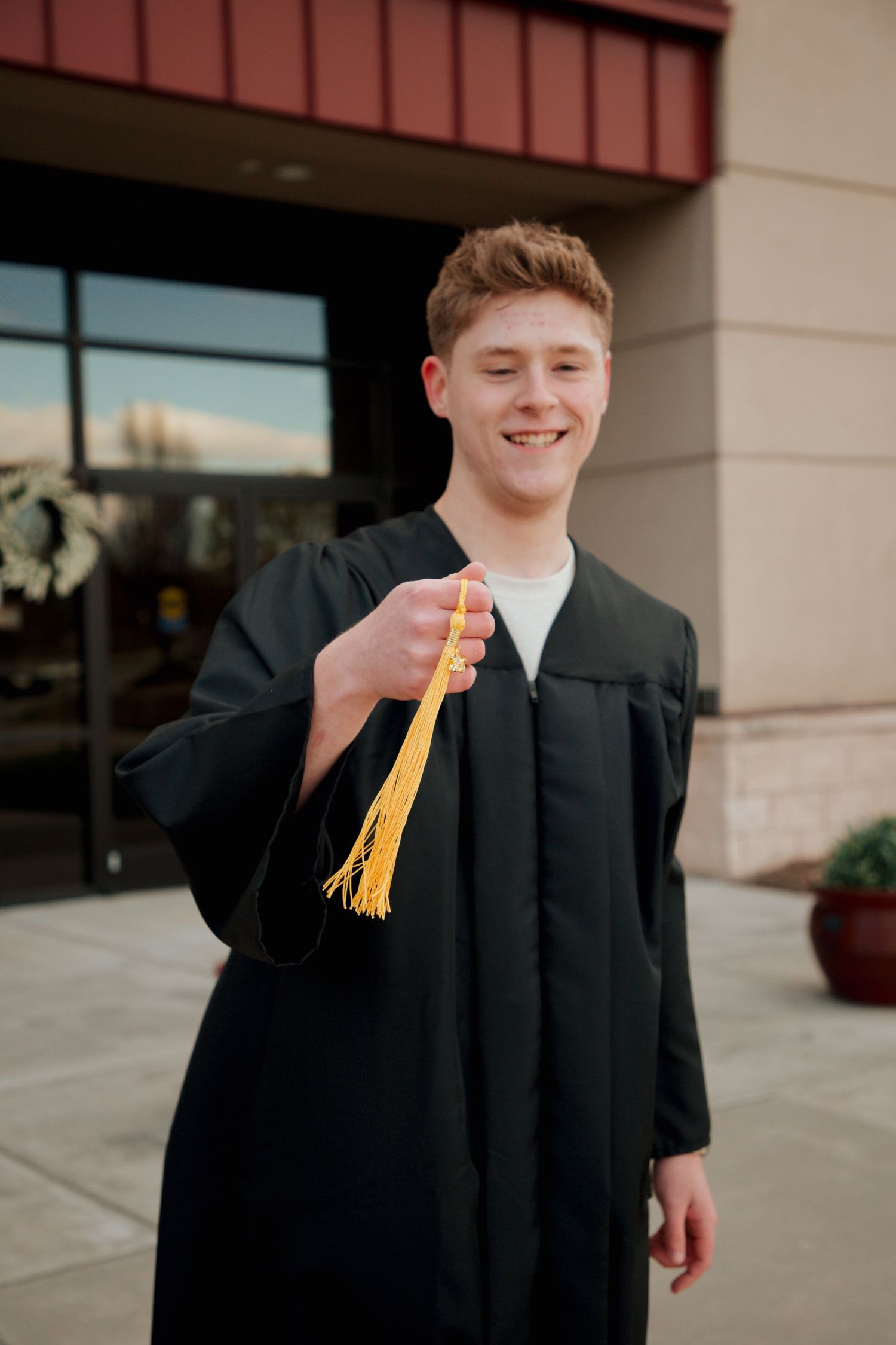 Young man in black graduation gown holding gold tassel toward camera outside school.