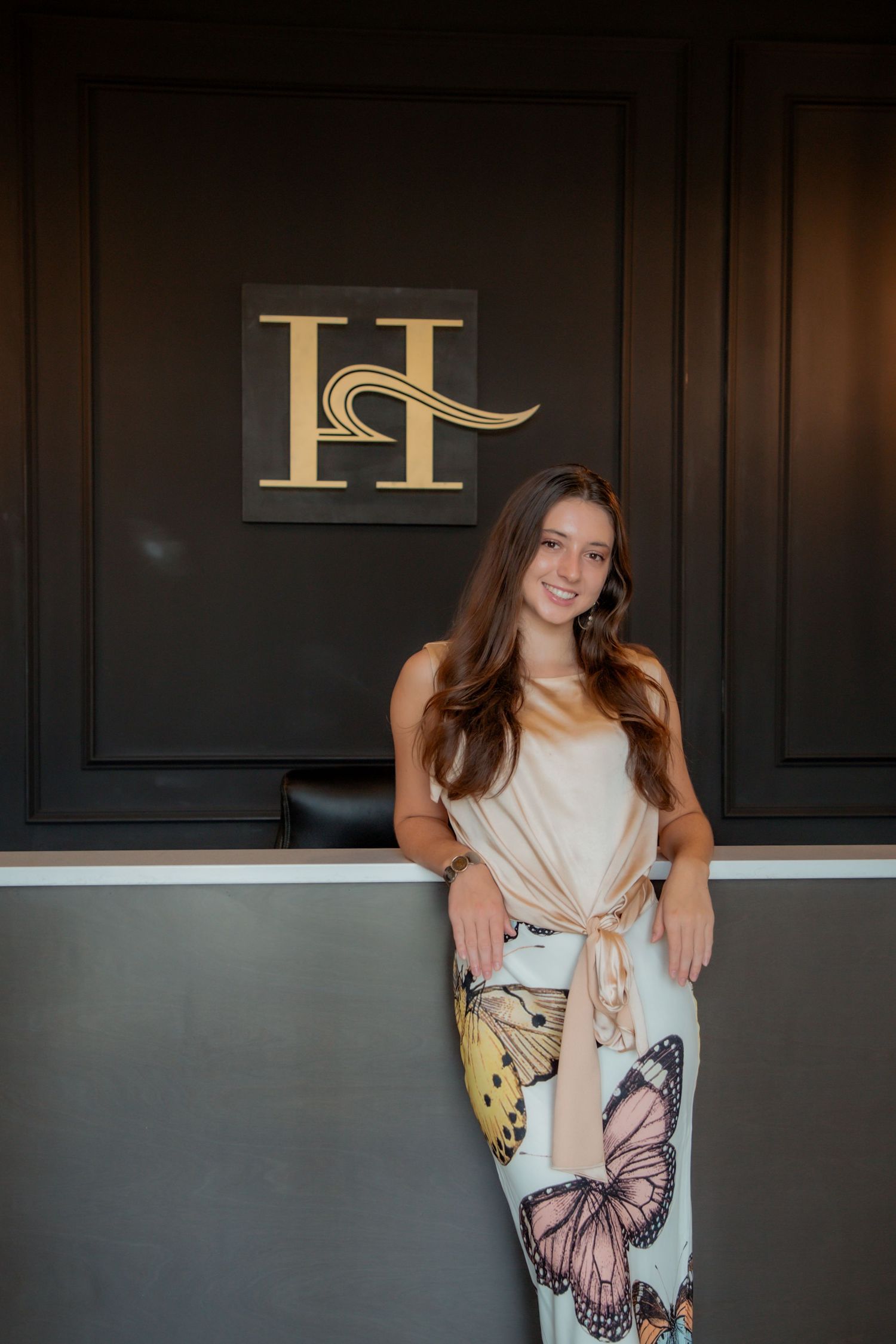 Smiling young woman in butterfly print skirt leaning against reception desk below gold H monogram.