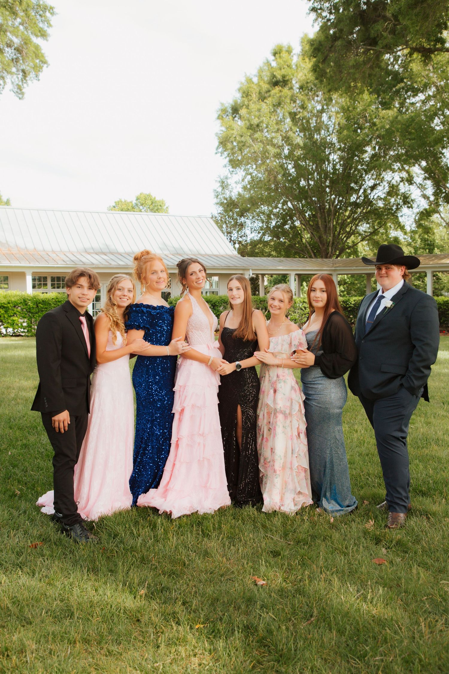 Group of teens in formal attire posing outdoors on grass for prom photos.