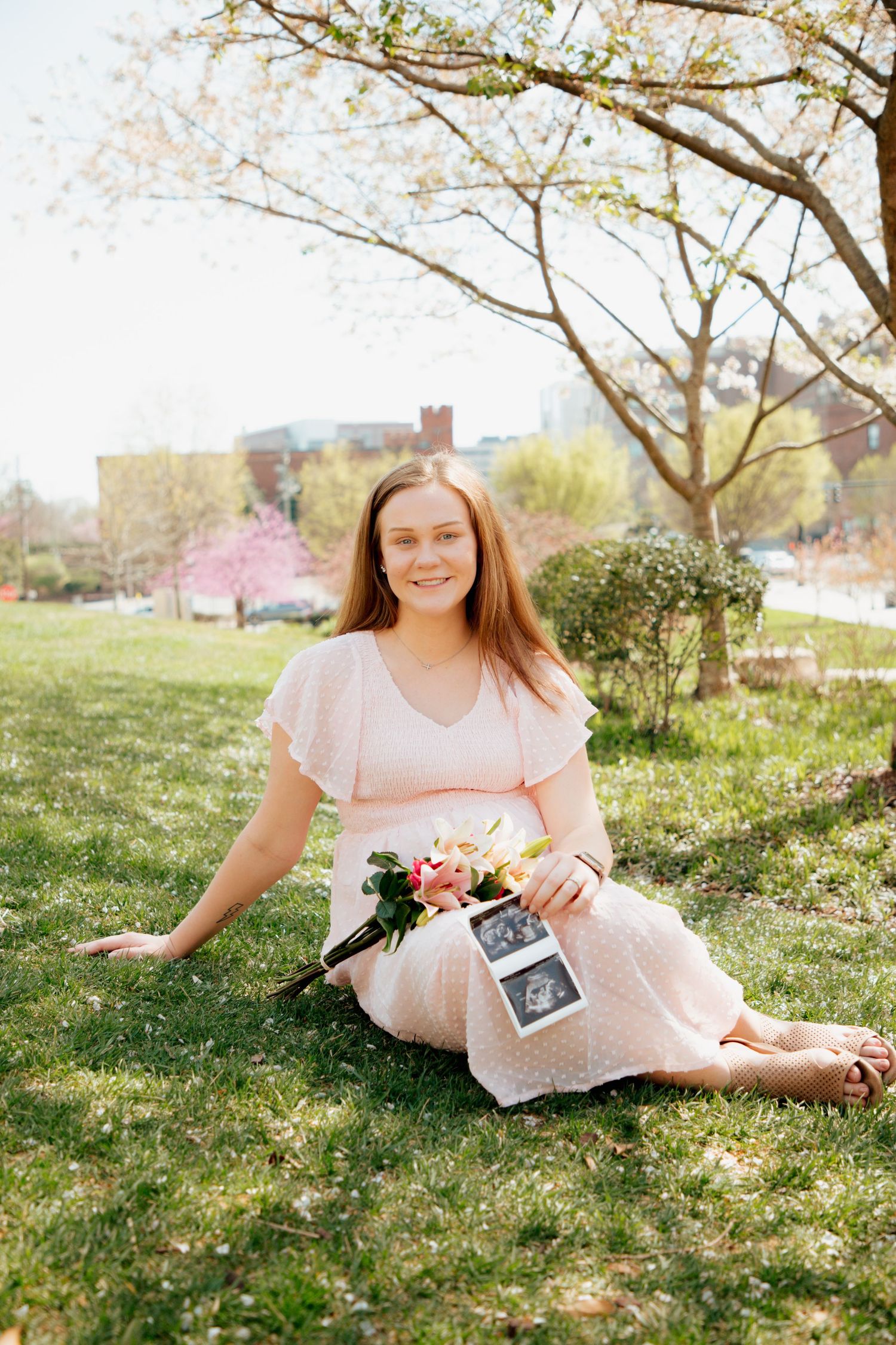 Pregnant woman in pink dress sitting on grass holding flowers and ultrasound photos.