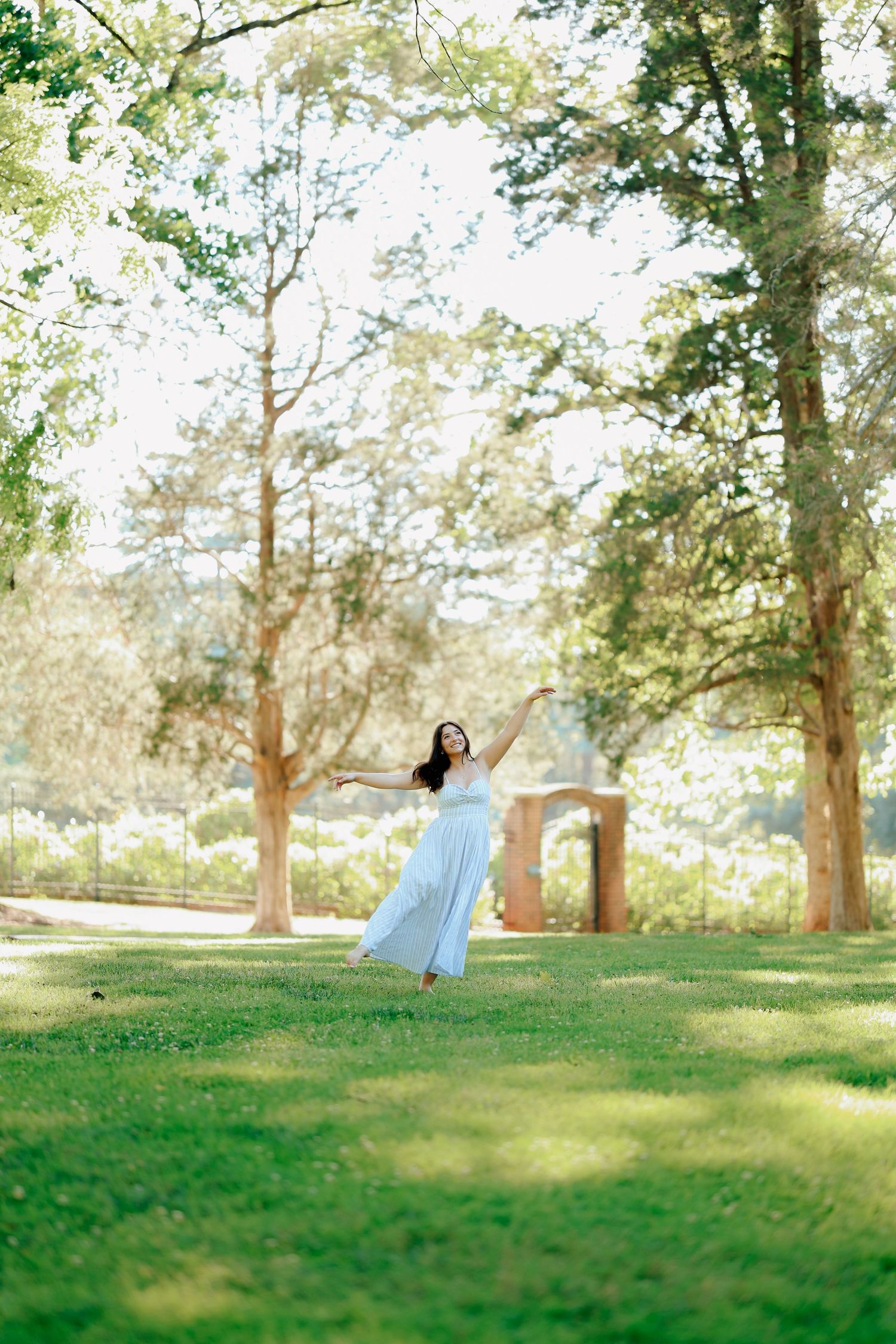 Woman in a white maxi dress with arms outstretched standing in a sunny green park with tall trees.