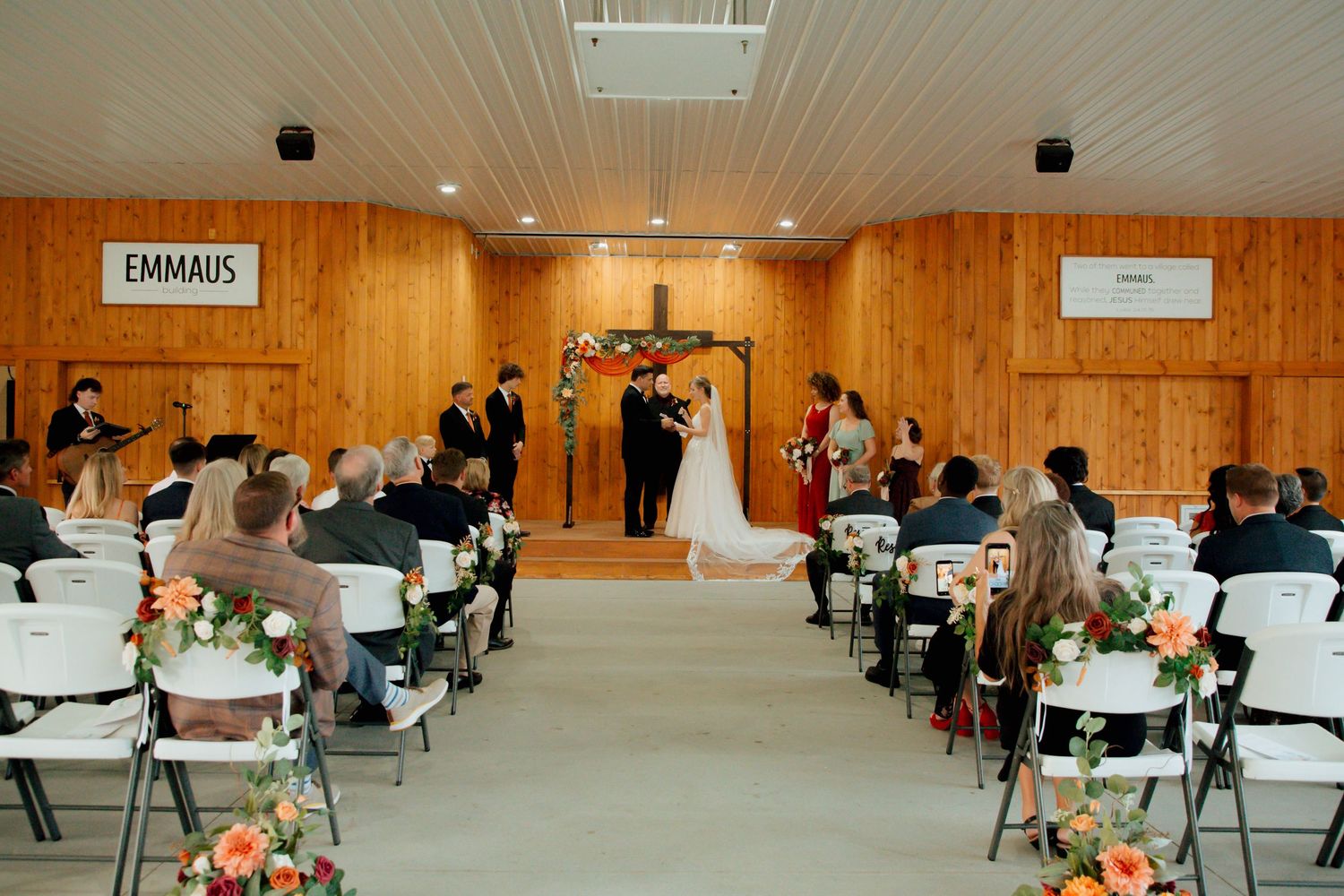 Wedding ceremony at Emmaus chapel with guests seated in white chairs along floral-lined aisle.