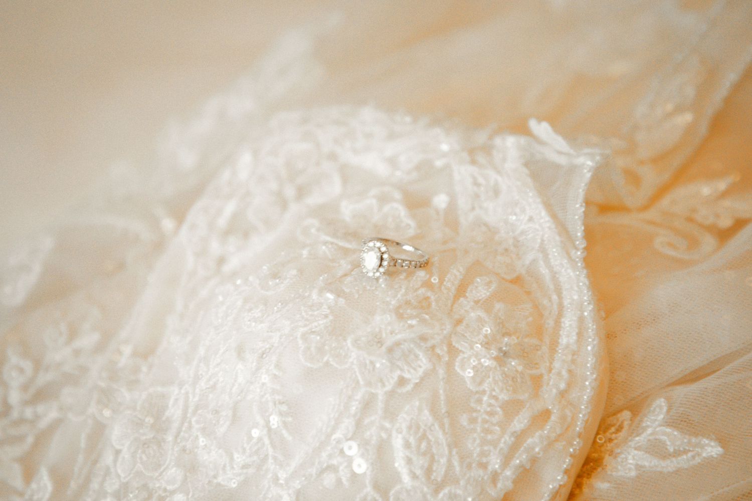 Close-up of a diamond solitaire engagement ring placed on delicate white lace wedding gown fabric.