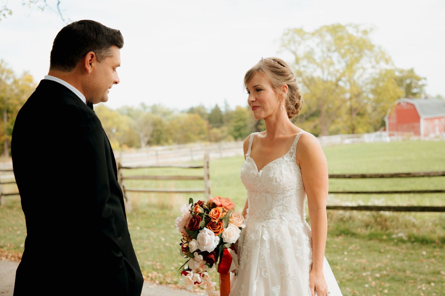 Bride holding autumn bouquet gazing at groom near red barn at fall wedding venue.