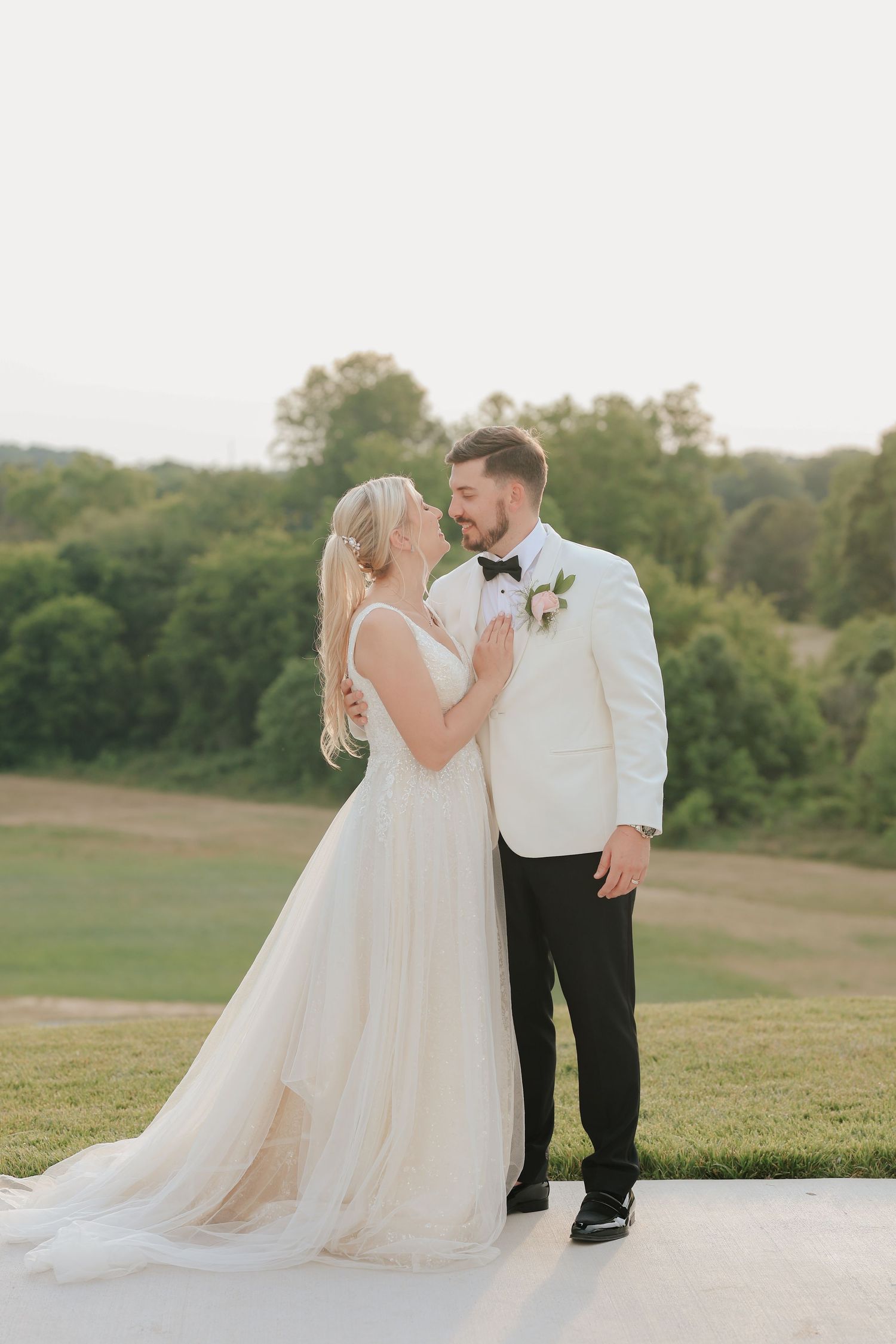 Bride and groom share a tender moment at golden hour on lush green hillside.