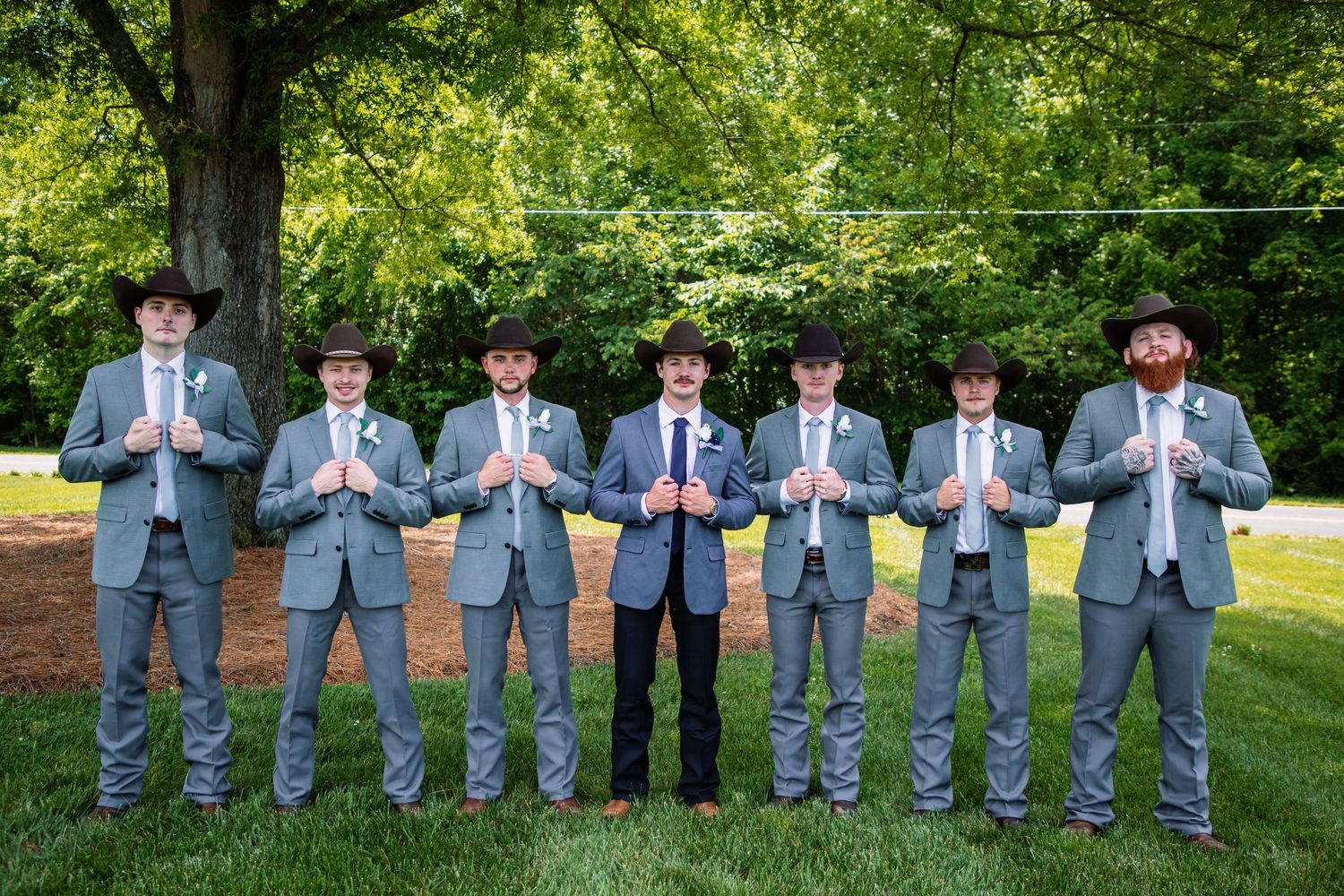 Seven groomsmen in matching gray suits and cowboy hats pose in a line at an outdoor wedding celebration.