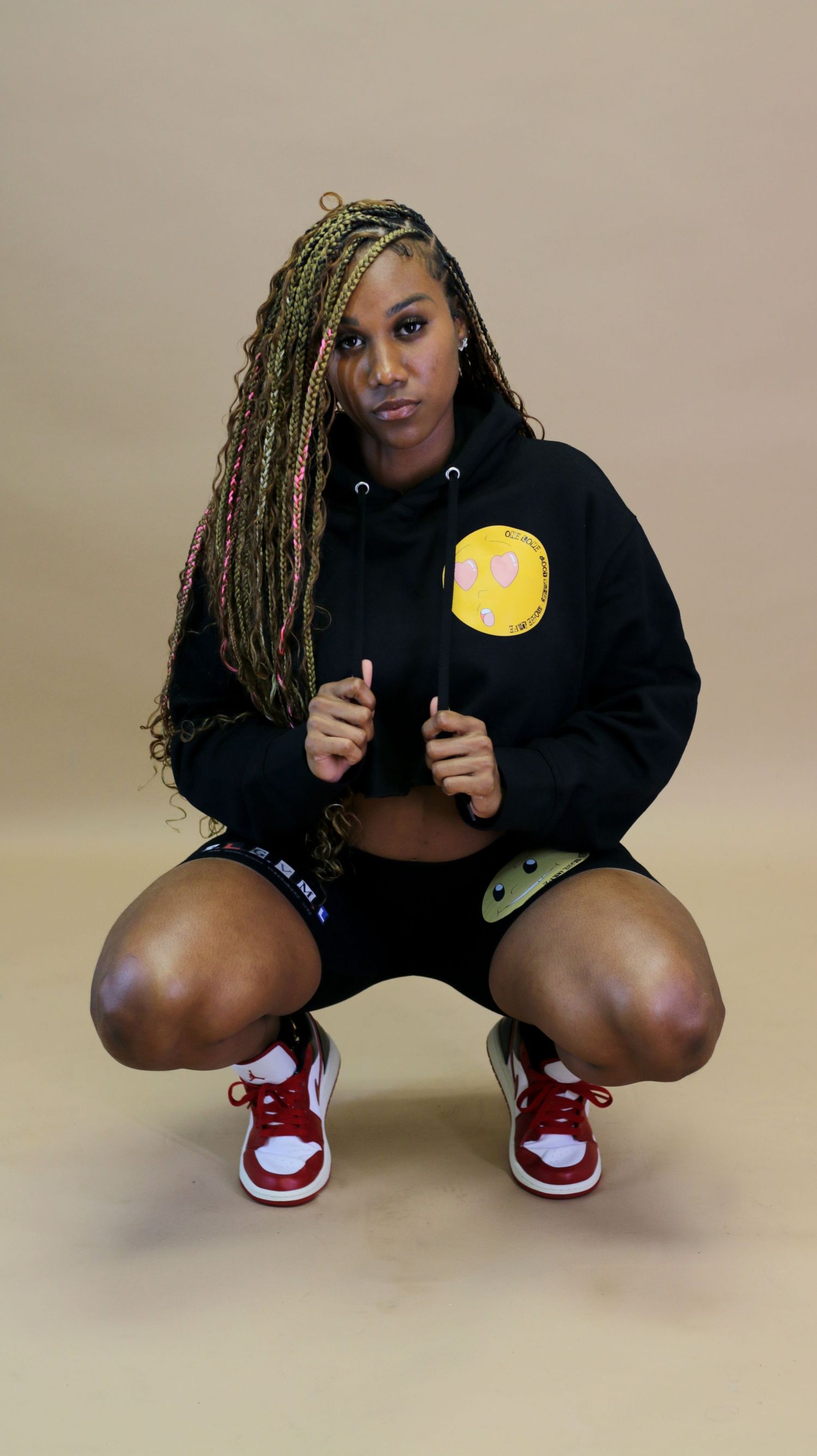 Woman with colorful braids in black hoodie and shorts squats wearing red Jordan sneakers.